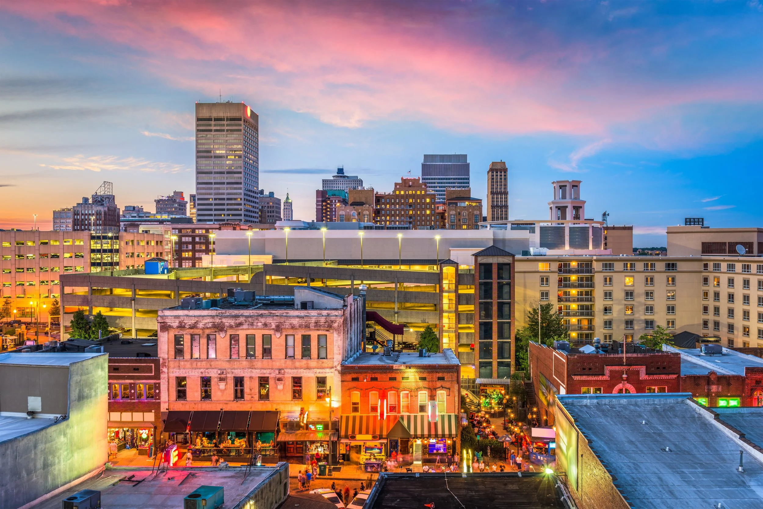 A city skyline during sunset with tall buildings and a busy street level filled with shops and people.
