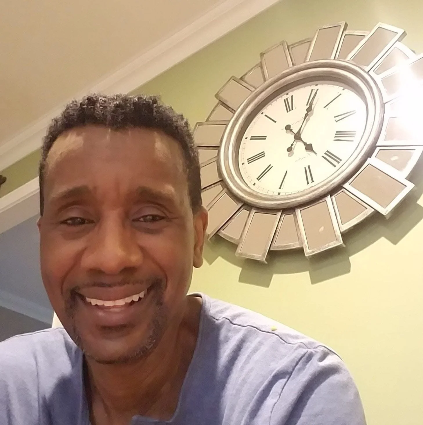 A smiling man with short curly hair taking a selfie in front of a large wall clock with Roman numerals.