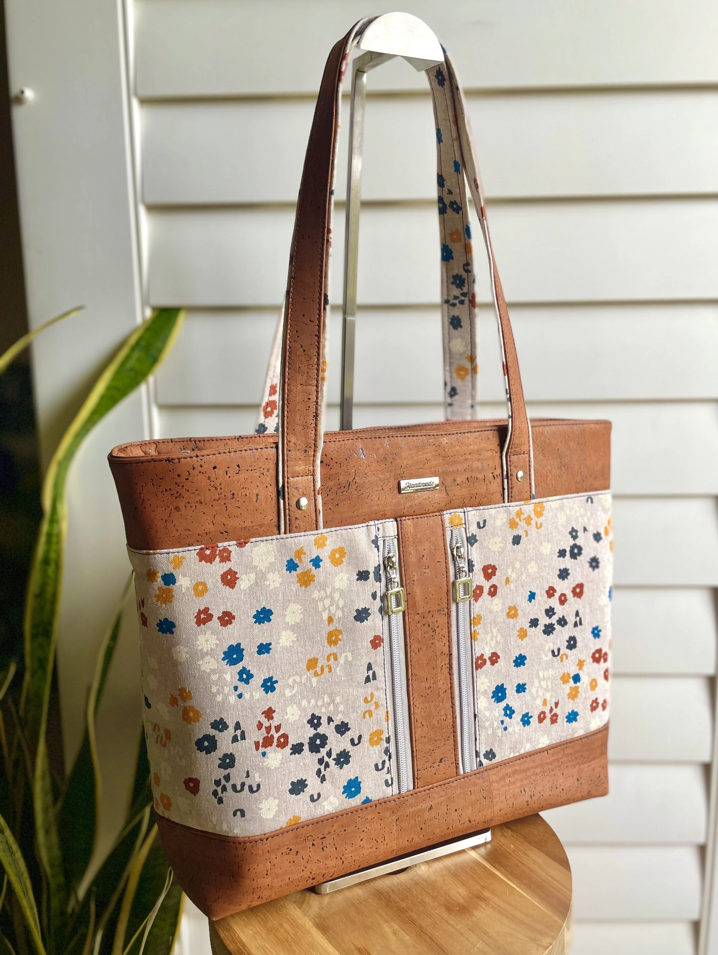 A tote bag made of cork and fabric with a multicolored floral pattern and front zipper pockets, sitting on a wooden stool.