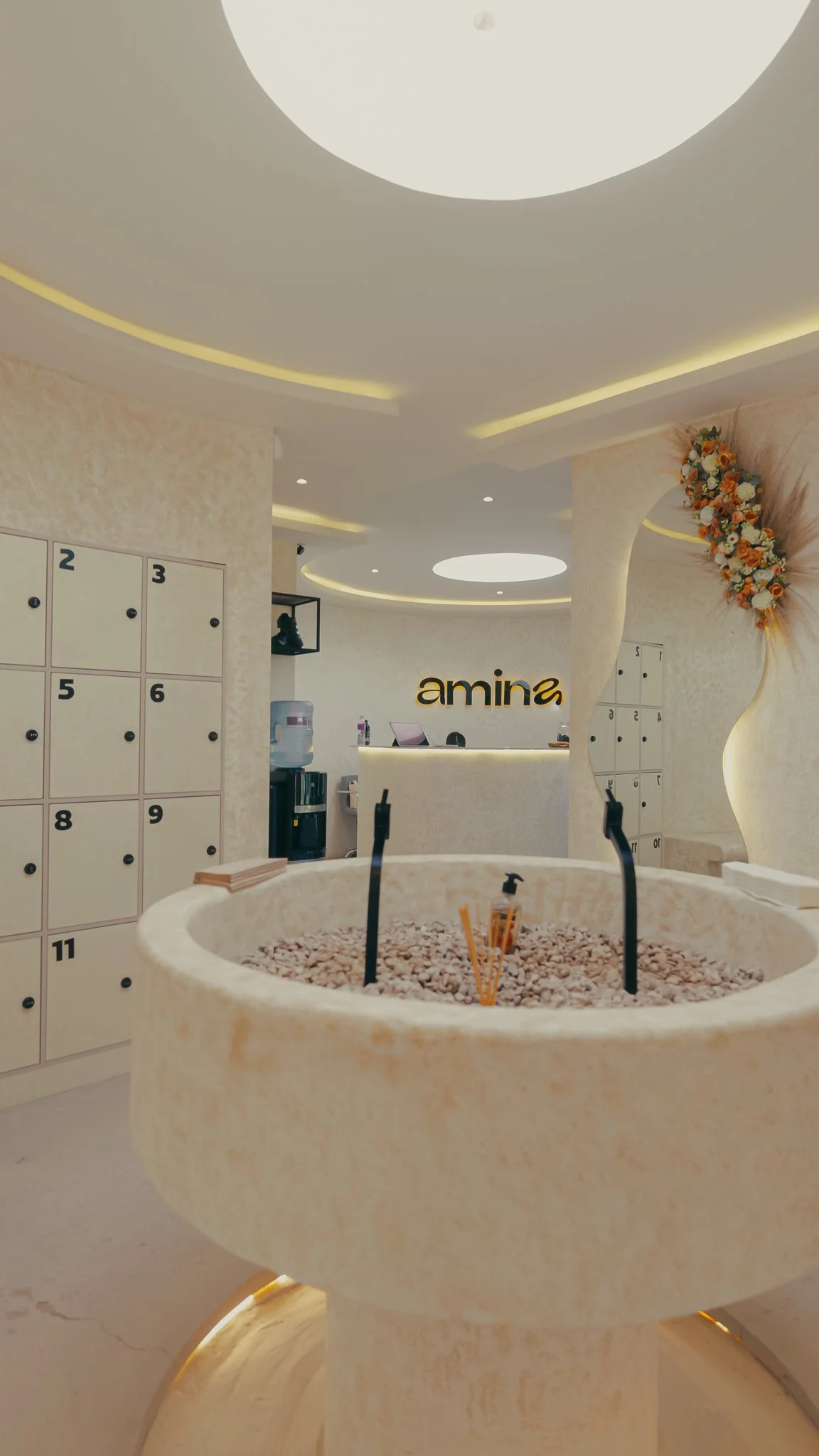 Recepción de estudio de ciclismo centro de bienestar, con lockers numerados, una decoración floral, una mesa de piedra con agua y accesorios, y un cartel que dice 'amina'.
