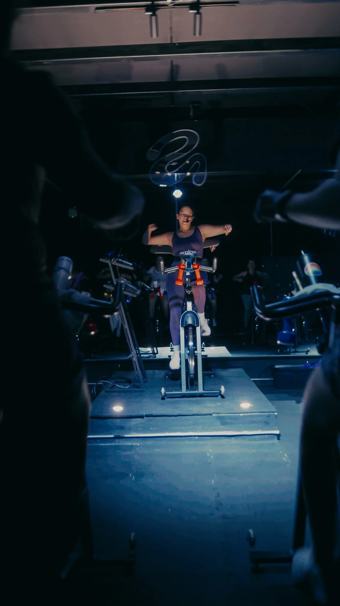 Una mujer participando en una clase de spinning en un gimnasio, montando en una bicicleta estática bajo luces azules, con otras bicicletas y participantes en el fondo.