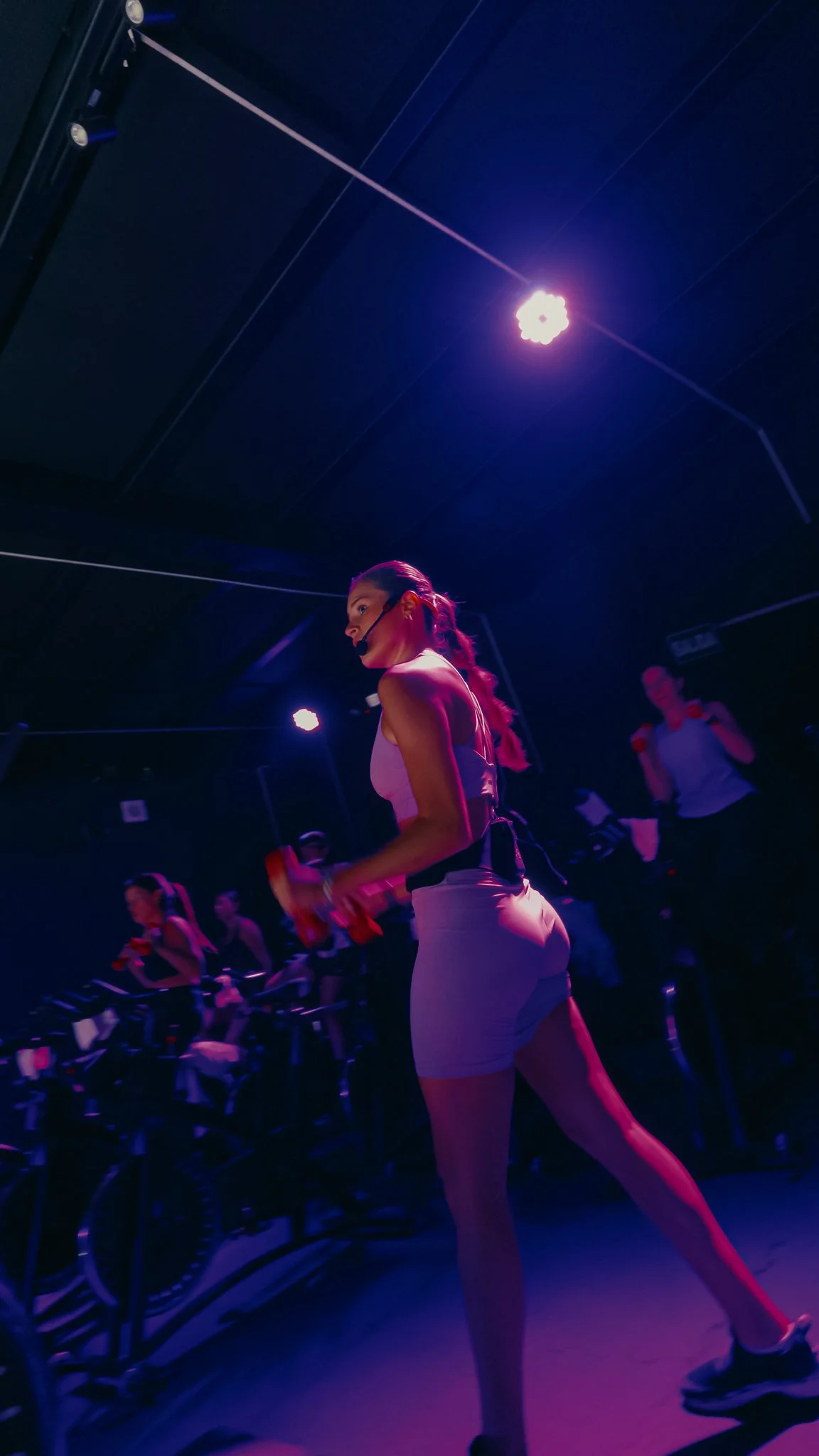 Mujer en clase de spinning en un estudio con iluminación morada y negra, usando un micrófono en la cabeza y sujetando unas manijas de bicicleta estática.