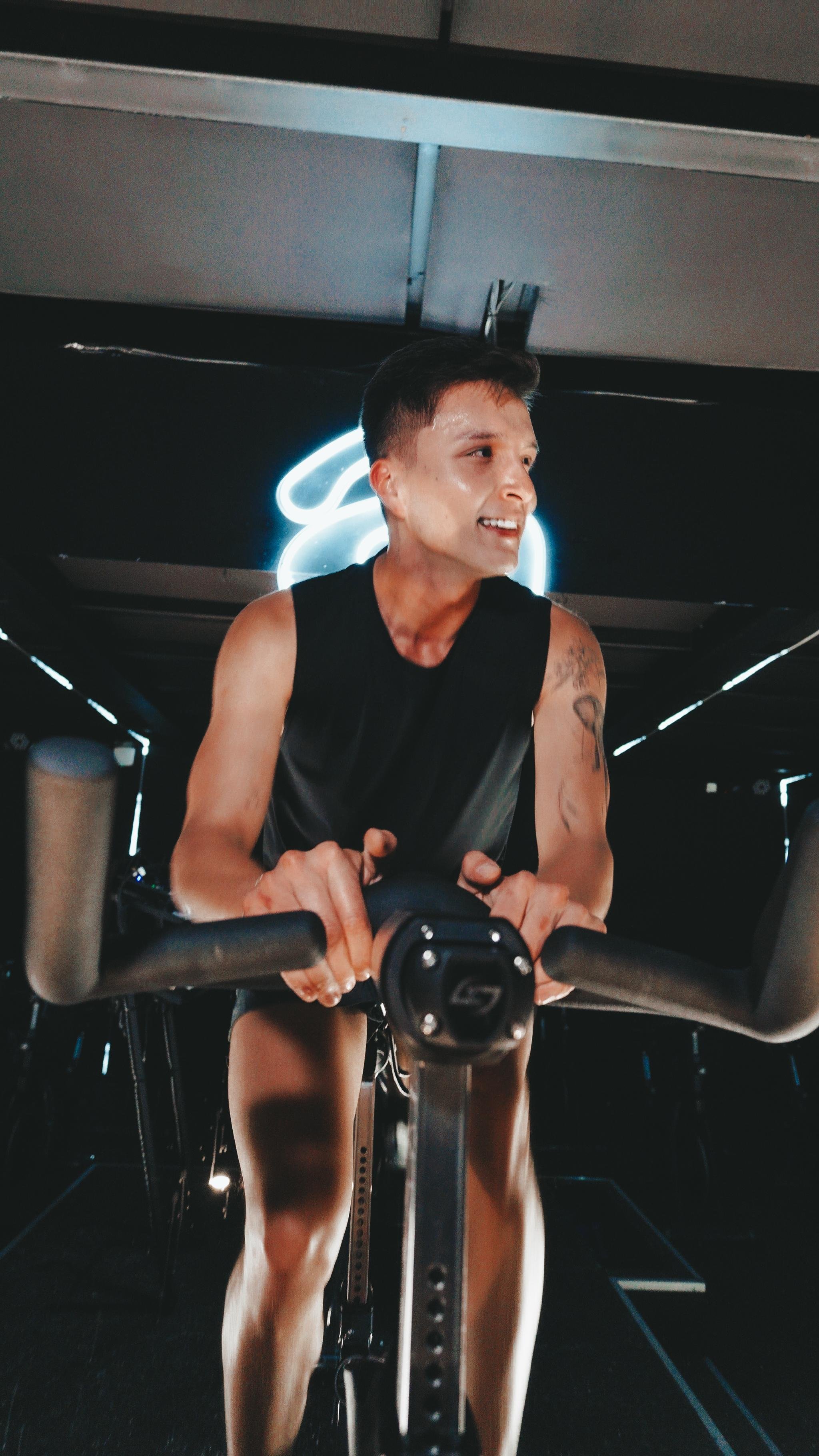 Persona haciendo ejercicio en una bicicleta de spinning en un gimnasio con iluminación tenue y fondo oscuro.