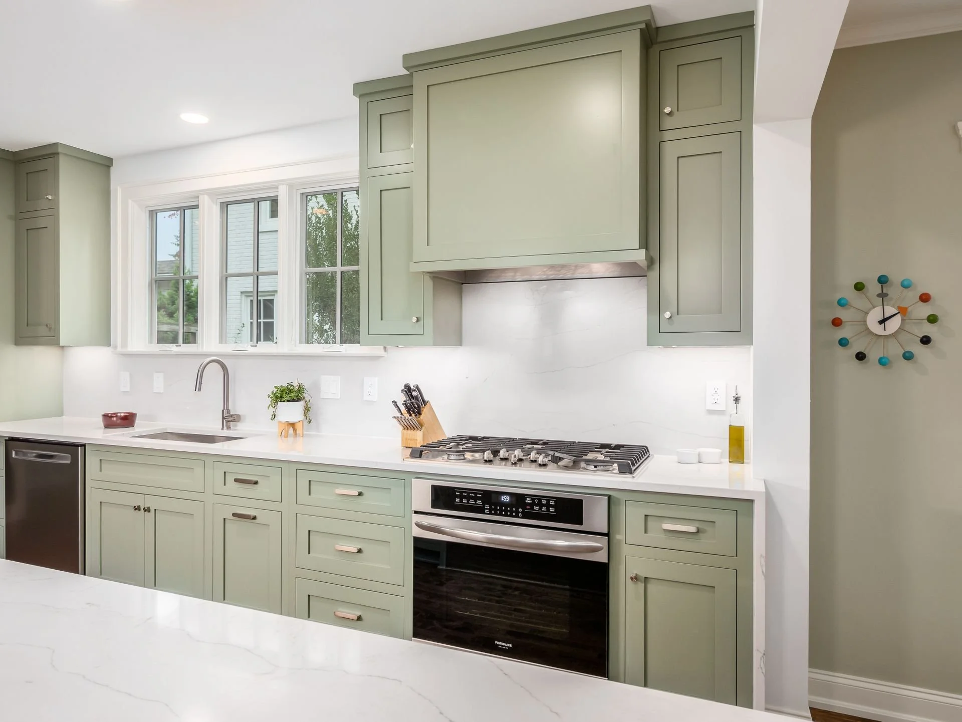 Kitchen with green cabinets, white marble countertops, and a window above the sink. Contains a built-in oven, stovetop, knife block, small potted plant, and decorative clock on the wall.
