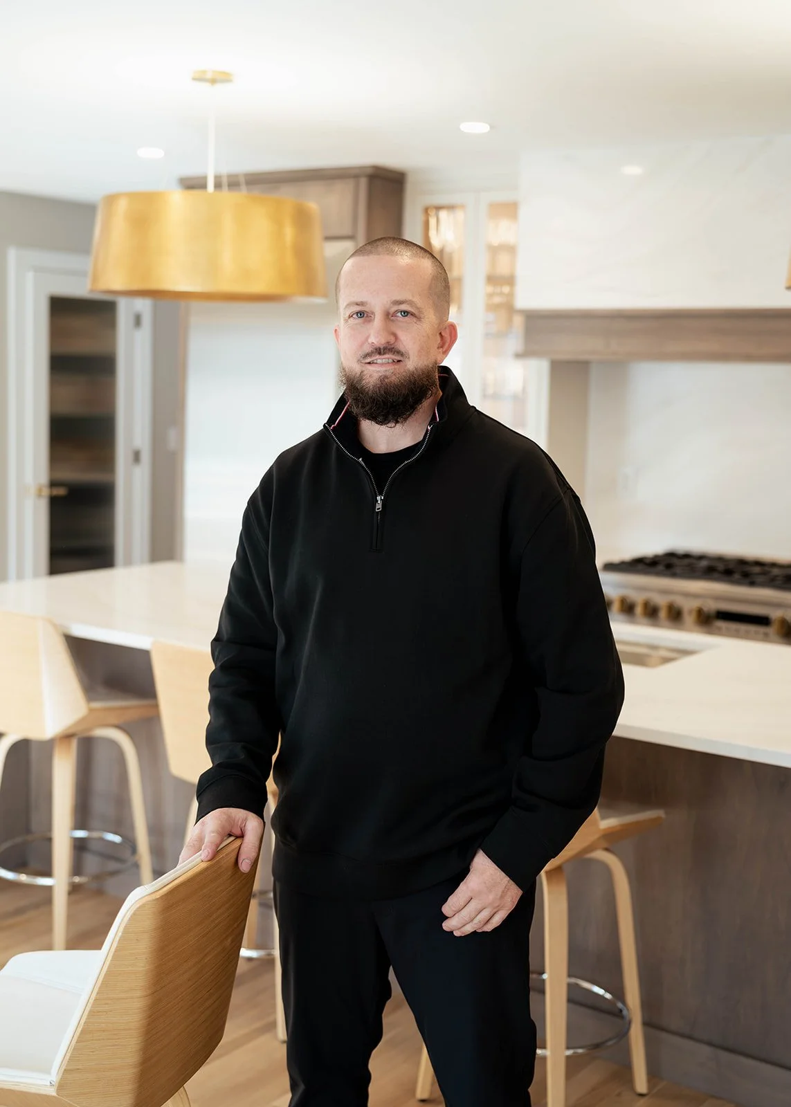 A man with a beard standing in a modern kitchen, resting his hand on a wooden chair.