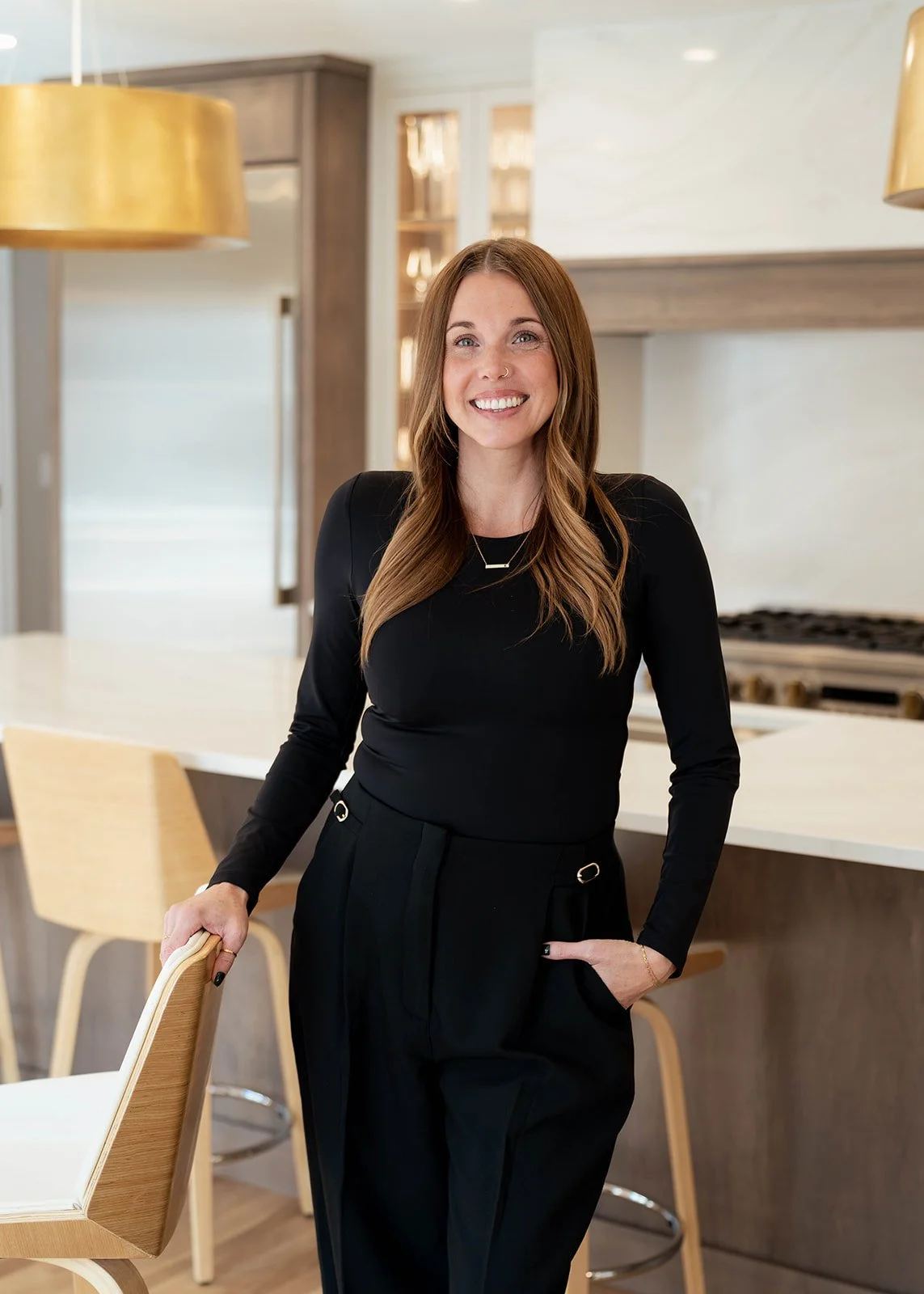 Woman in black outfit standing in modern kitchen, smiling, with a countertop and chairs in the background.