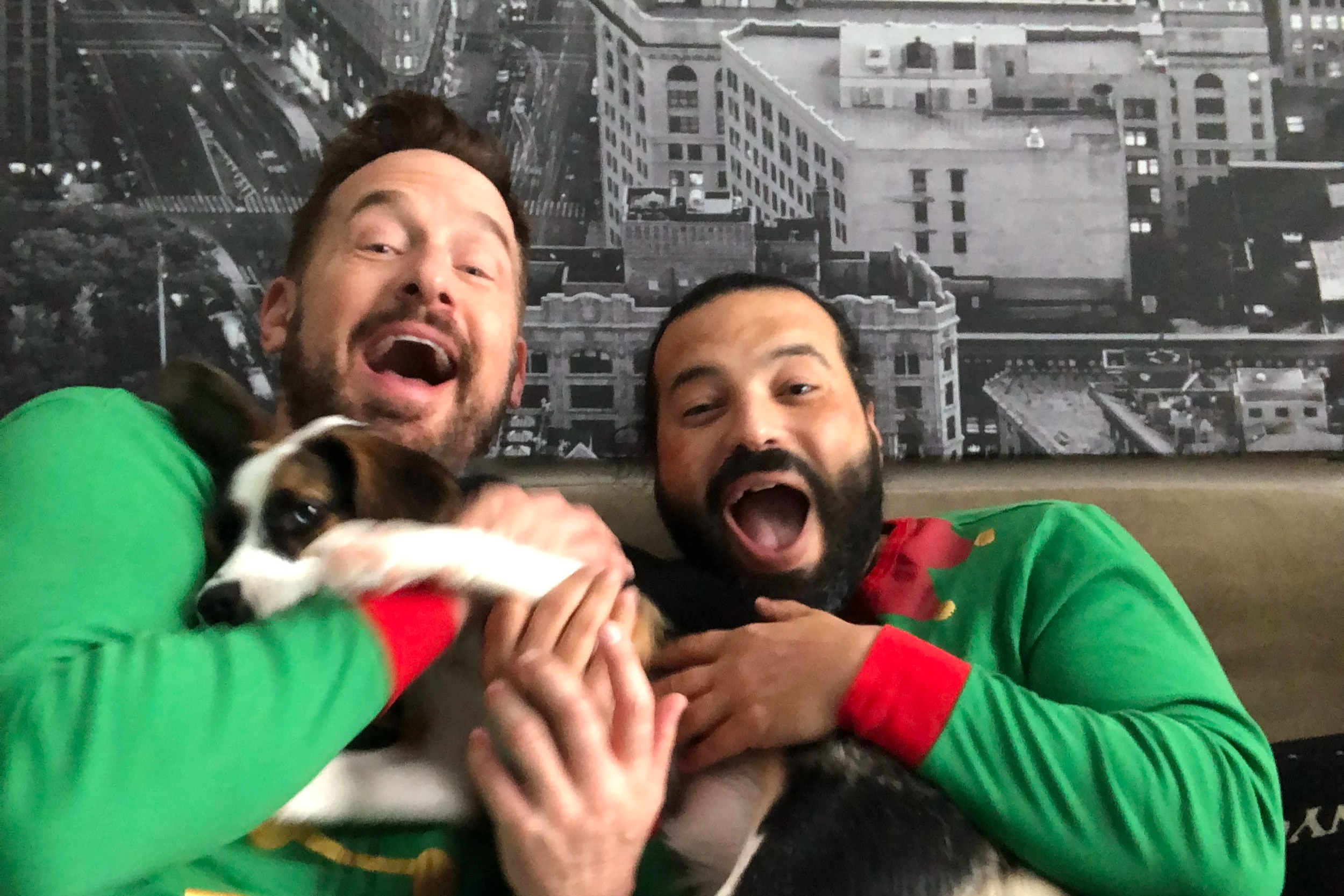 Two men wearing Christmas sweaters smiling and holding a puppy, with a cityscape mural in the background.