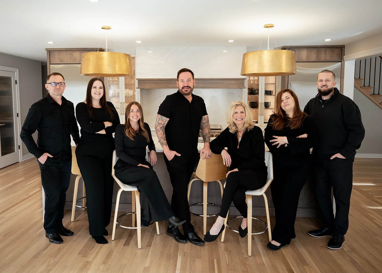 Group of seven people in black clothing posing in a modern kitchen with wooden flooring and pendant lights.