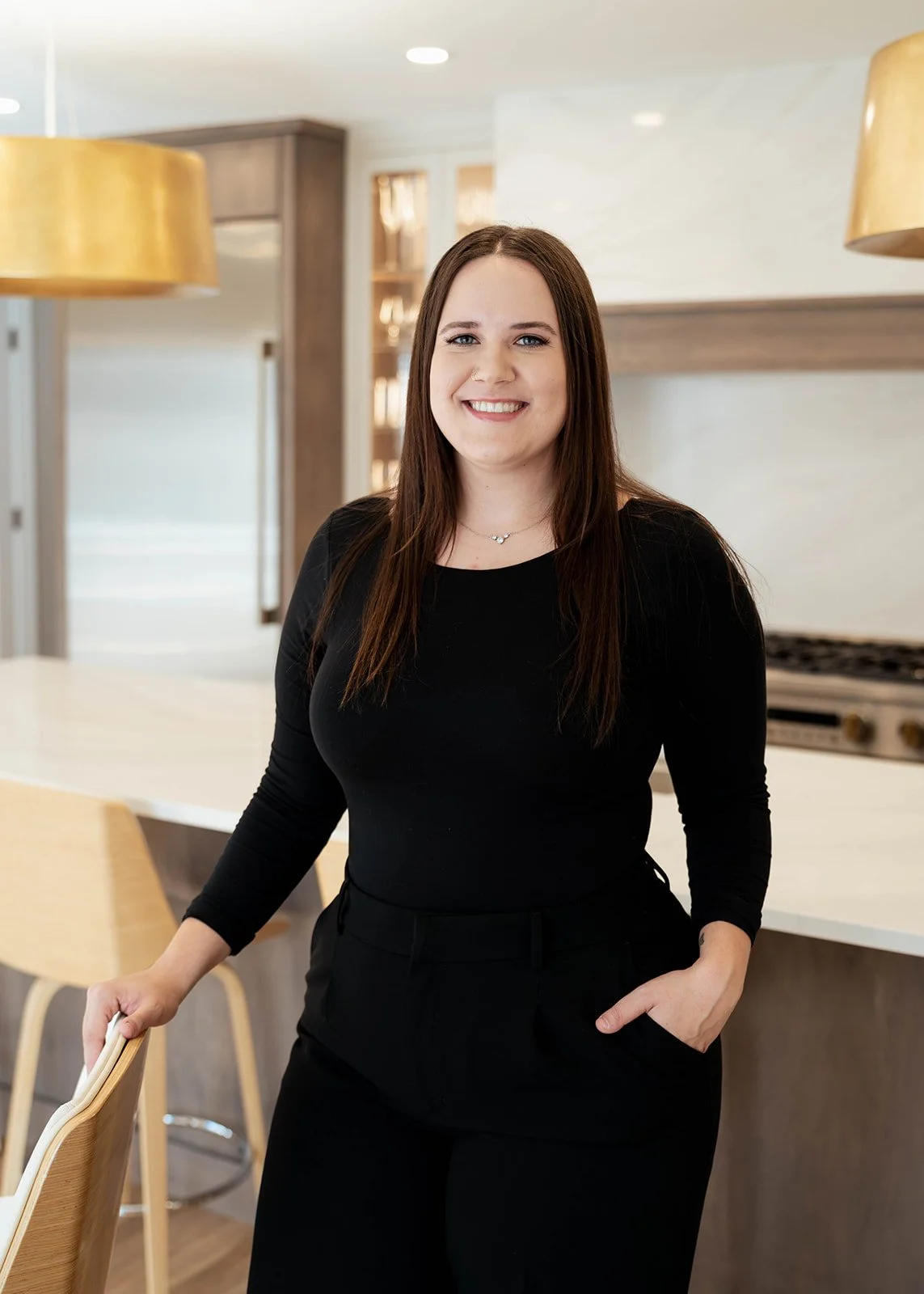 A woman with long brown hair wearing a black long-sleeve top and black pants, smiling and standing in a modern kitchen.