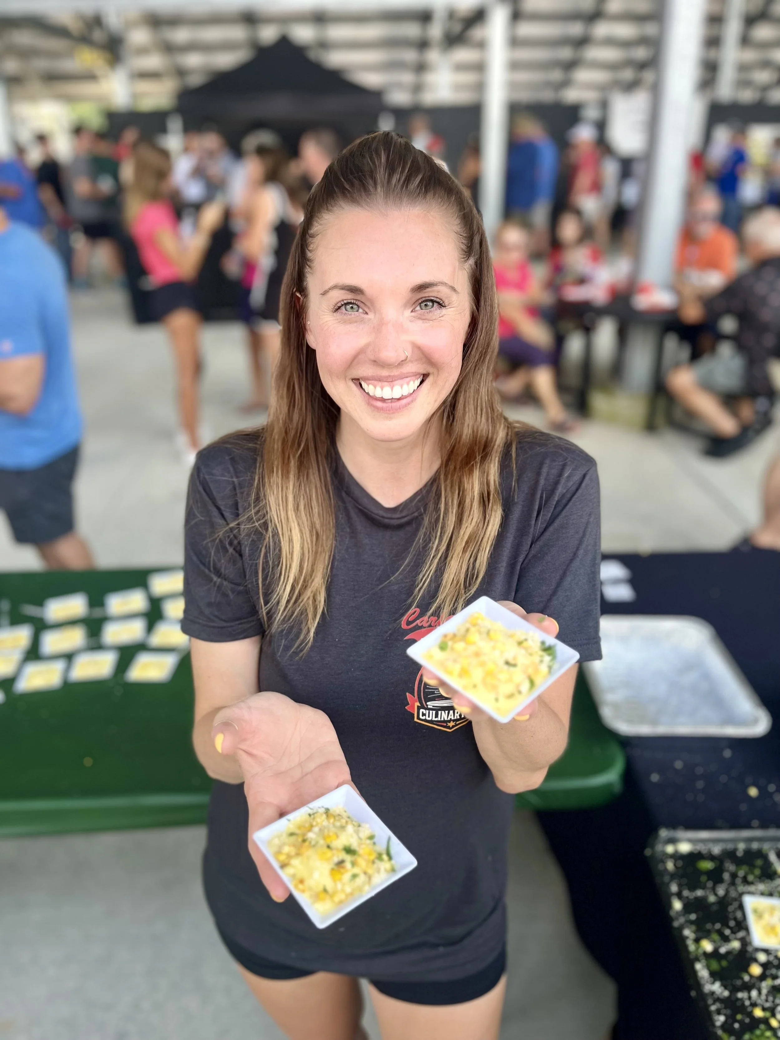 A smiling woman with long brown hair holding two small bowls of food at a crowded outdoor event.