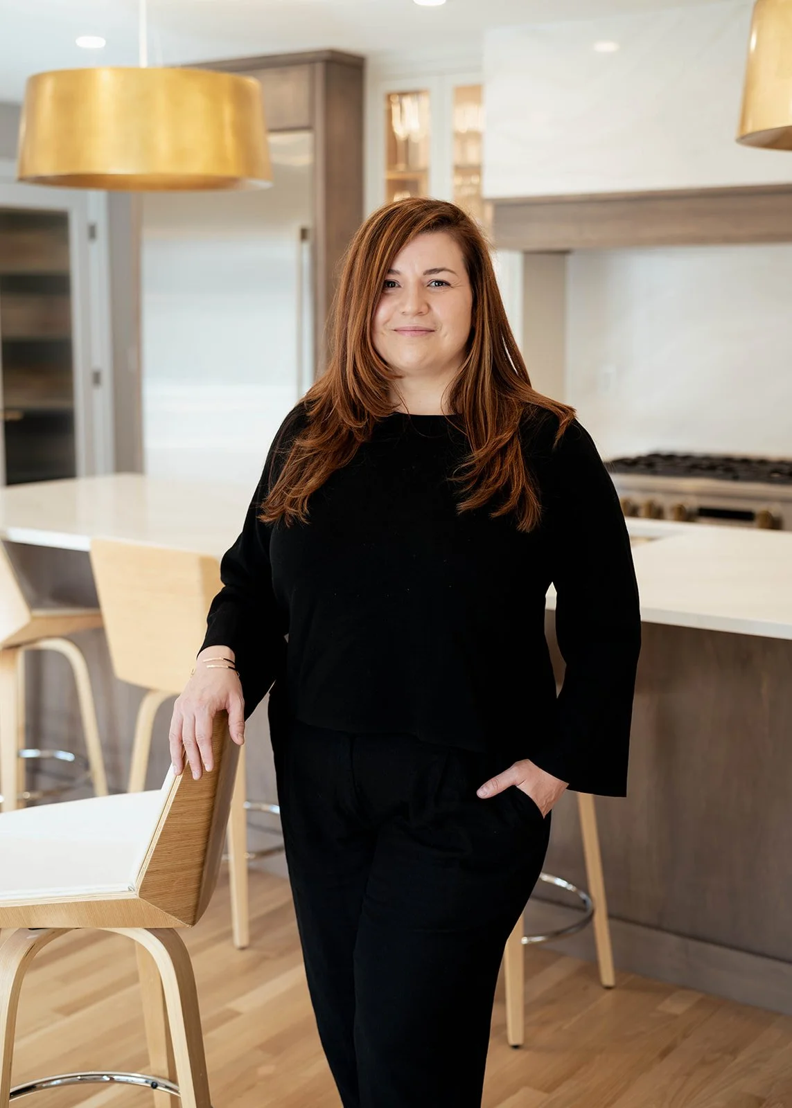 A woman with shoulder-length red hair wearing a black top and black pants, standing in a modern kitchen with wood and white accents. She is smiling slightly with her right arm resting on a high chair.