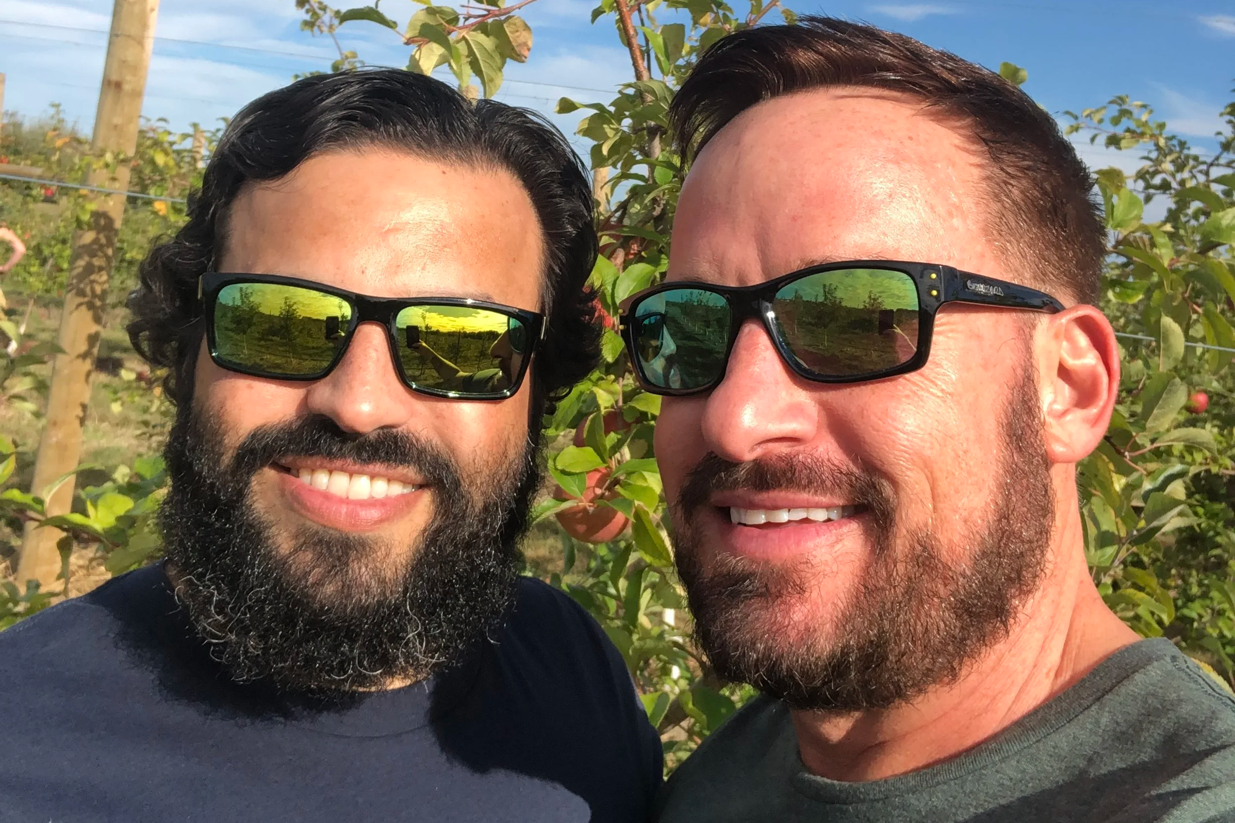 Two men smiling outdoors in a sunny orchard, both wearing sunglasses, with green trees and blue sky in the background.