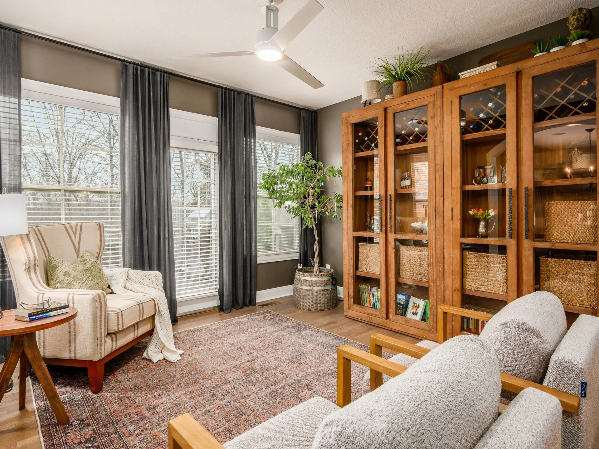 Living room with large windows, dark curtains, a wooden bookcase with glass doors, a potted plant, and a seating area with armchairs and a patterned rug.