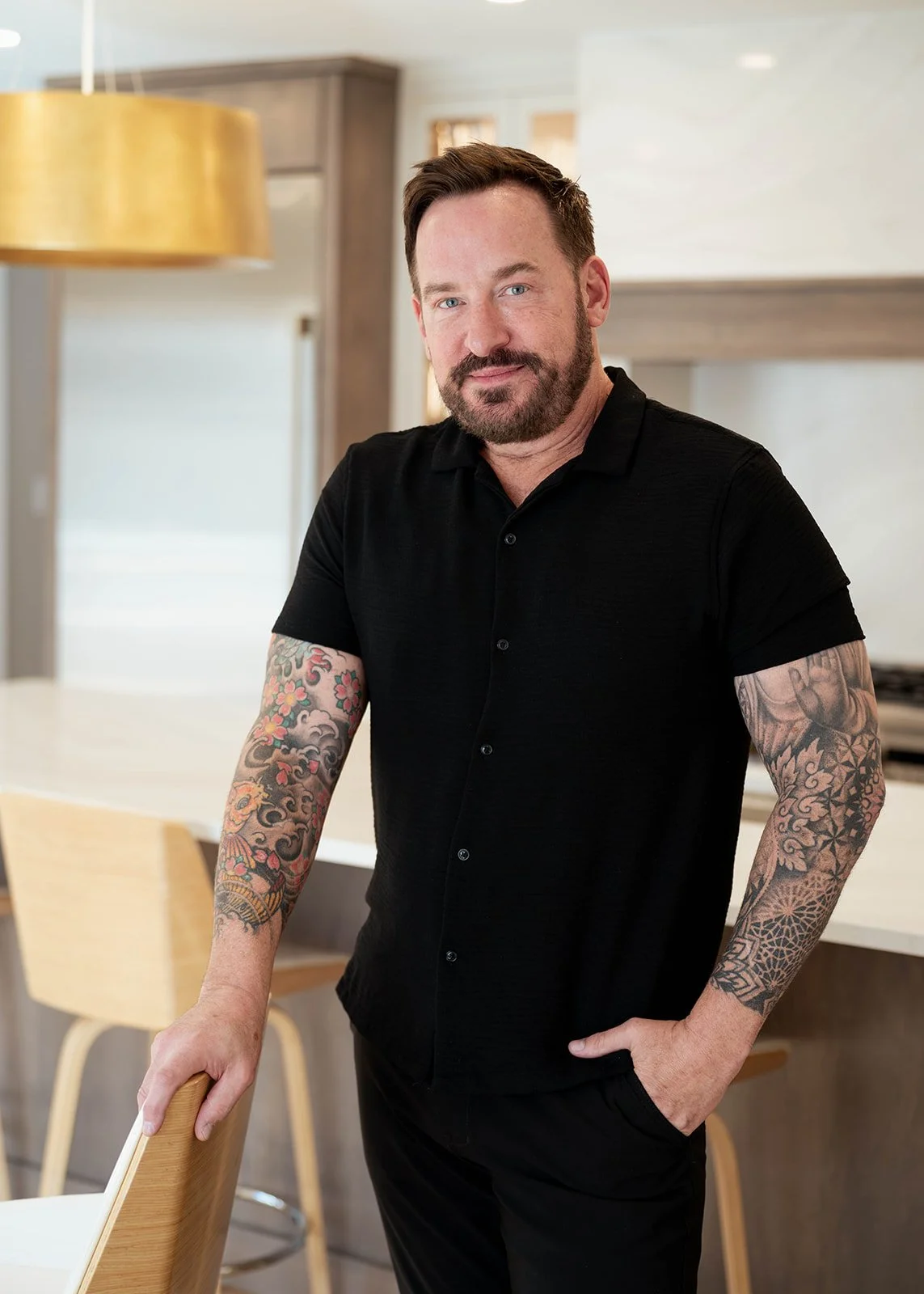 A man with tattoos on both arms, wearing a black shirt, standing in a modern kitchen and leaning on a chair.