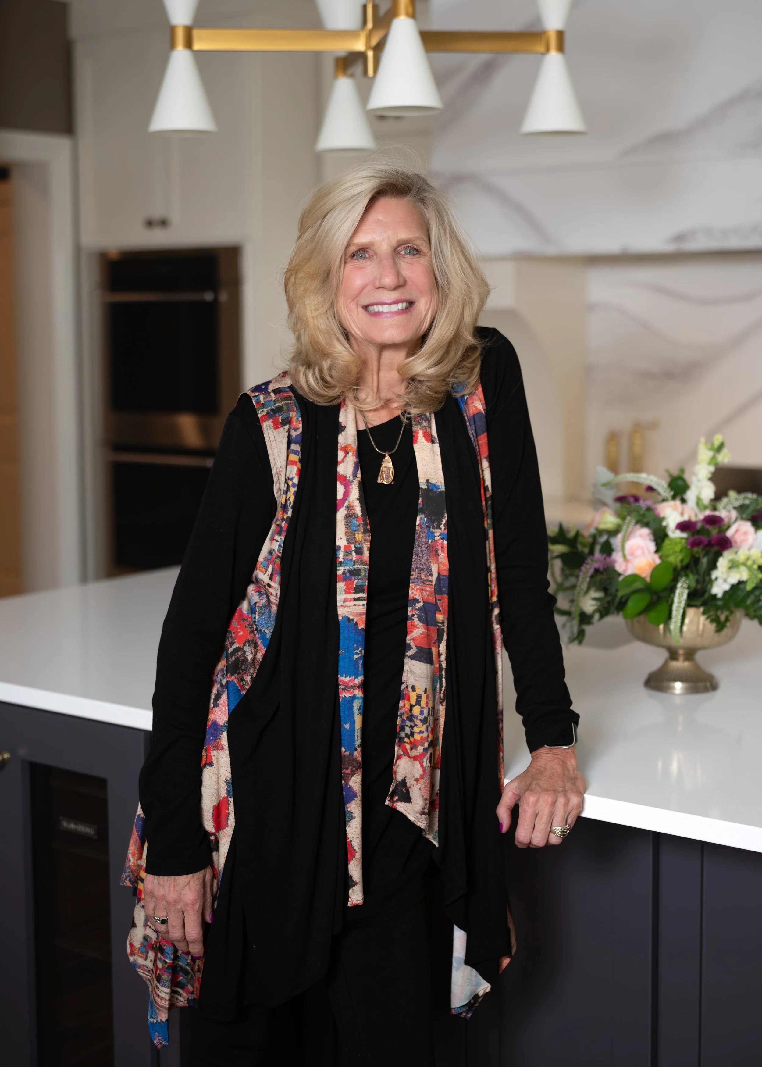An older woman with blonde hair, wearing a black dress and a multicolored scarf, stands in a modern kitchen next to a white countertop with a bouquet of flowers in a gold vase. Behind her are white cabinets and a black oven, with pendant lights hanging from the ceiling.