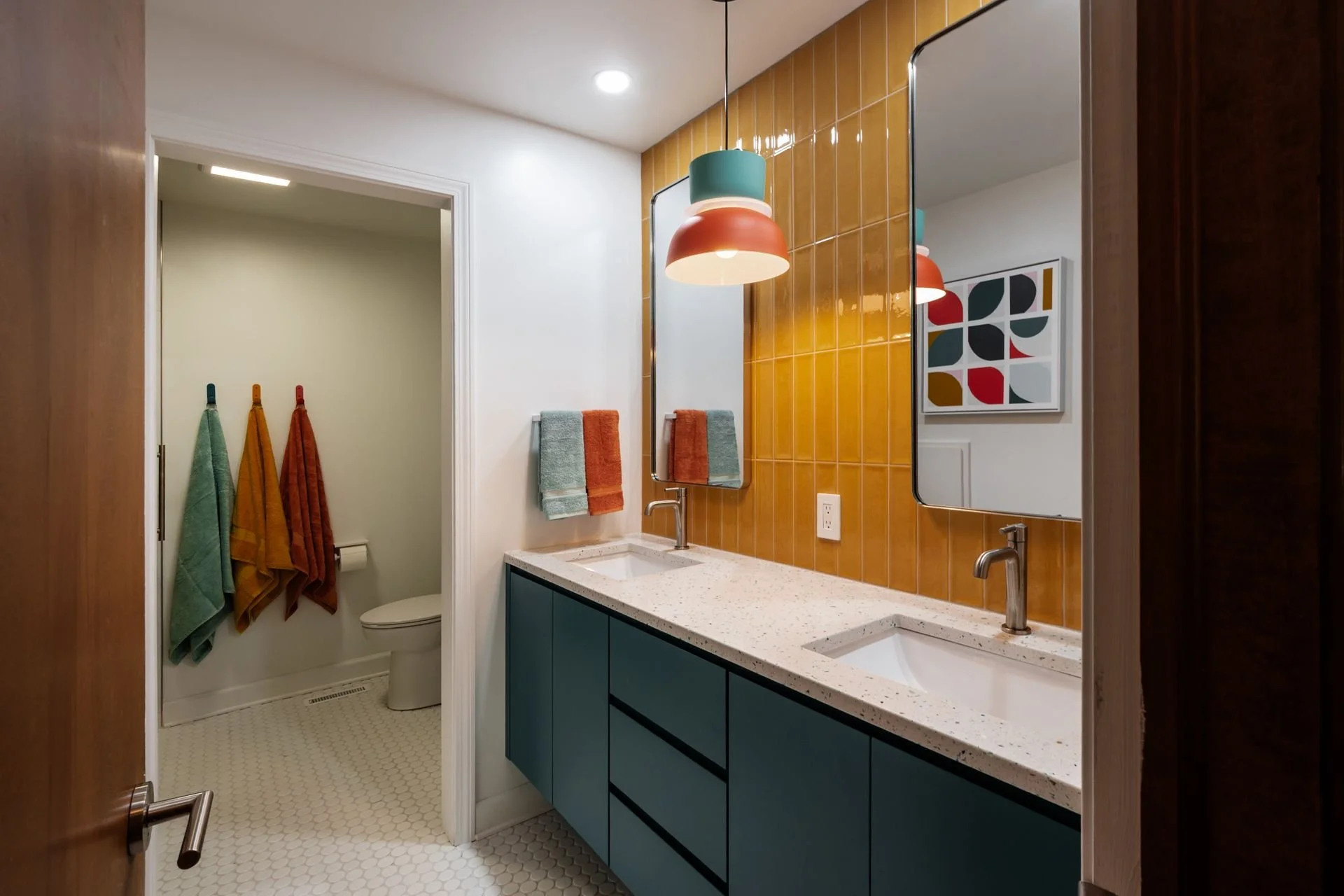 Bathroom with double blue vanity, terrazzo countertop, orange-backed yellow-tiled wall, and colorful pendant lights; seen through open wooden door, with a separate toilet area with hanging towels in the background.