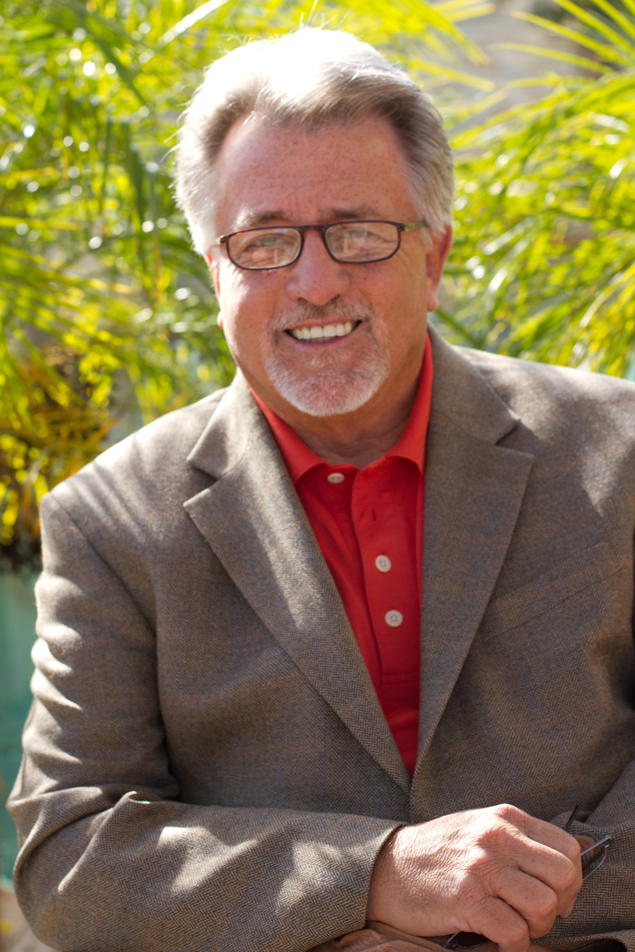 Portrait of a smiling older man with gray hair and beard, wearing glasses, a red shirt, and a tan blazer, standing outdoors with green foliage in the background.