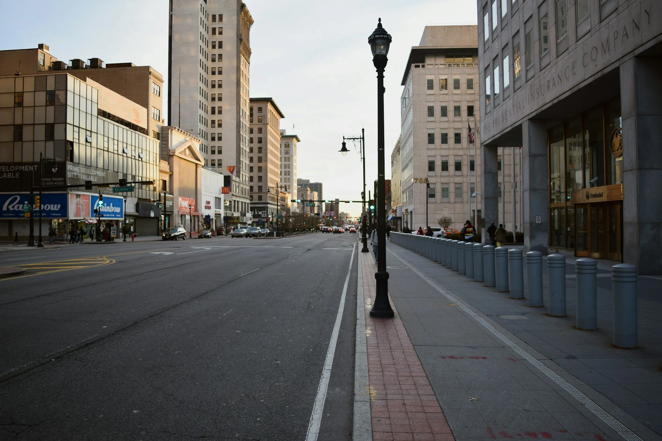 Empty city street with tall buildings, street lamps, and a row of bollards on the sidewalk during daytime.
