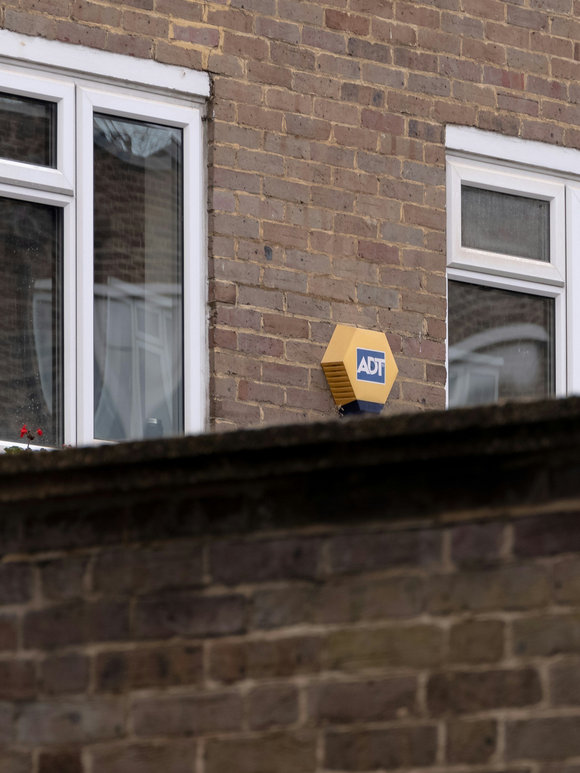 Photo of a brick building with two white-framed windows. A yellow ADT security alarm box is mounted on the wall between the windows. The photo appears to be taken from inside a vehicle, as part of the dashboard is visible at the bottom.