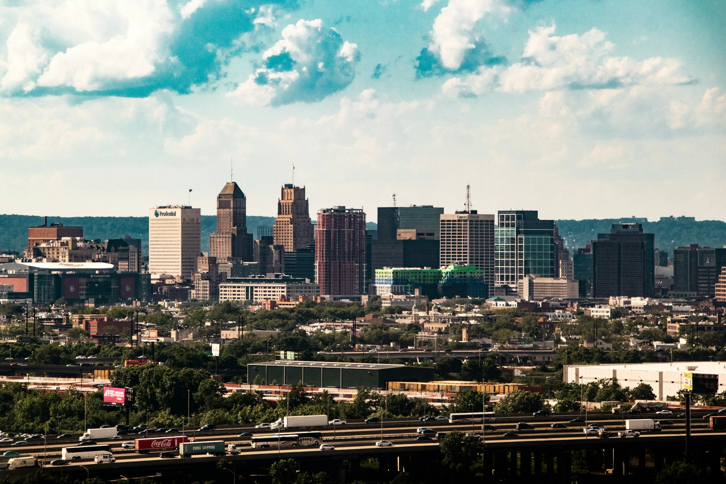 City skyline with tall skyscrapers, some having antennas or flagpoles, under a partly cloudy sky, with highway traffic and green areas in the foreground.