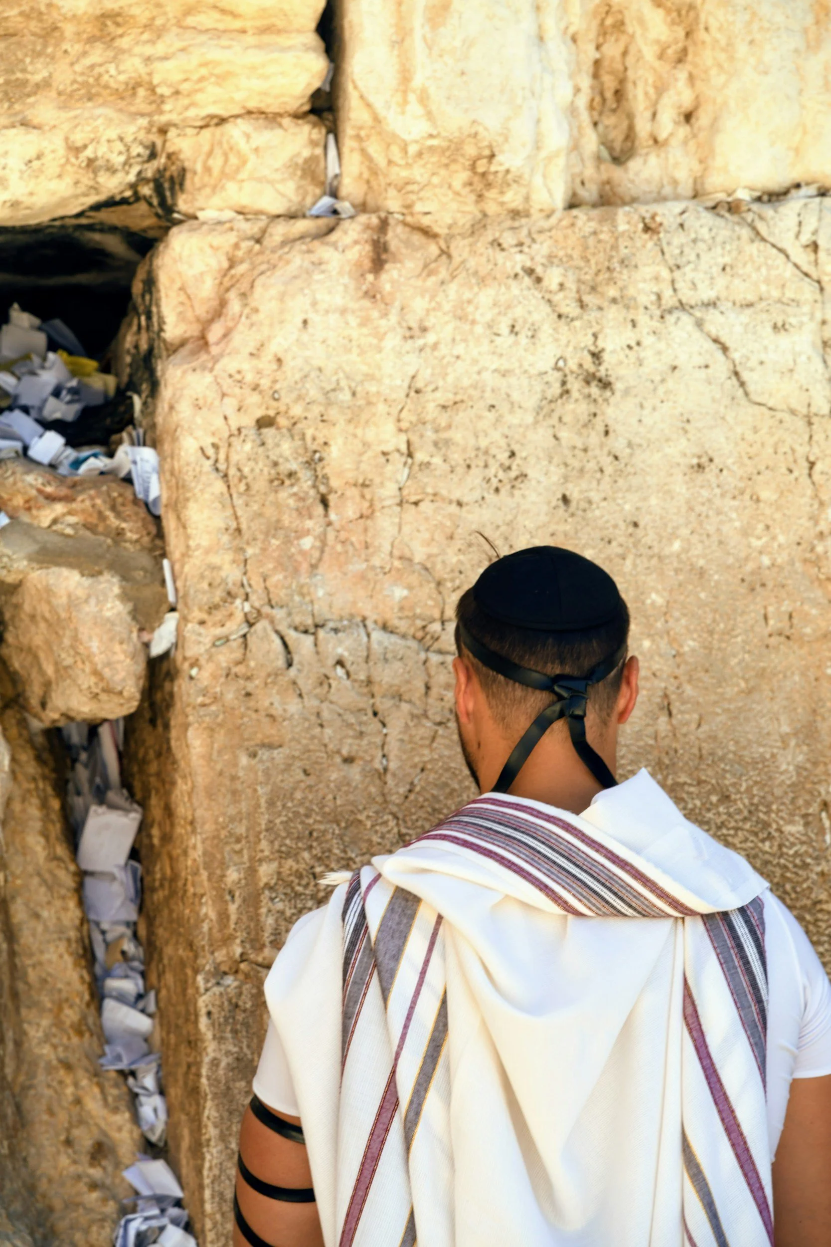 A man in traditional Jewish prayer attire, including a tallit and tefillin, standing in front of the Western Wall in Jerusalem.