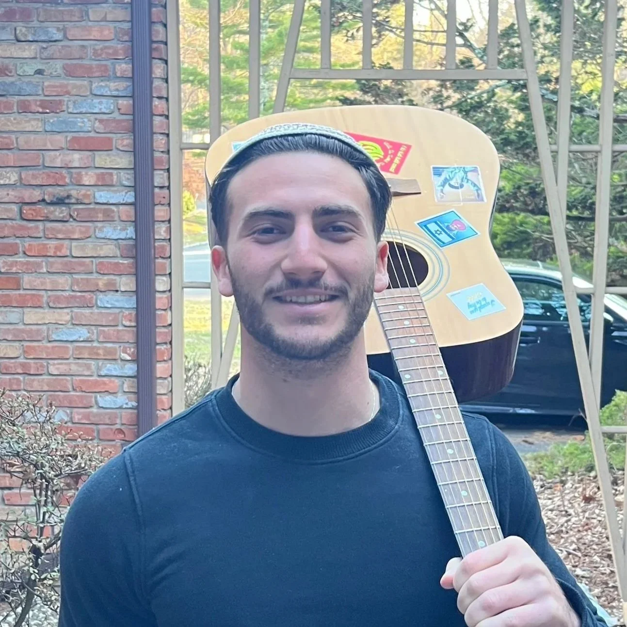 A young man smiles while holding an acoustic guitar over his shoulder outdoors, with a brick wall and a window reflecting greenery in the background.