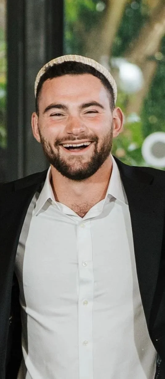 A man with a beard, smiling, wearing a white shirt, black blazer, and a light-colored cap, standing indoors with greenery visible outside through the window.