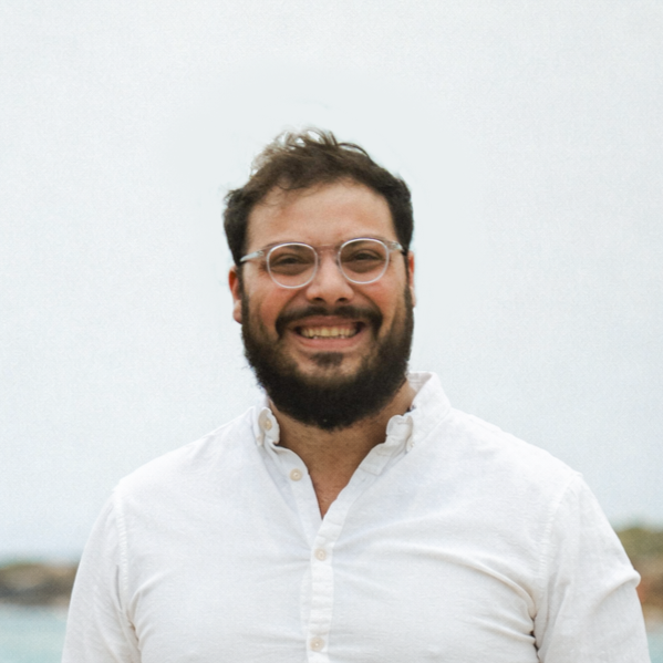 A smiling man with glasses and a beard wearing a white button-down shirt outdoors.
