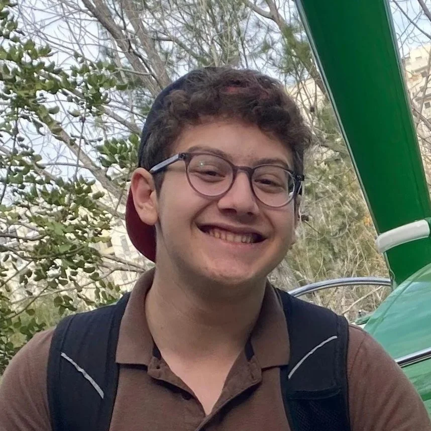 A young man with glasses smiling outdoors, wearing a cap backwards and a backpack.