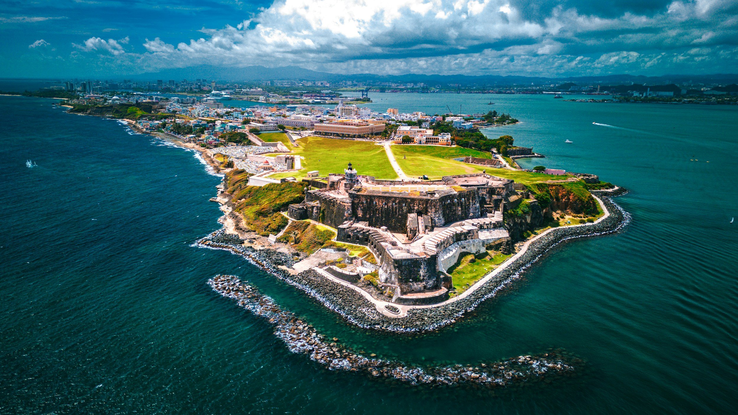 Aerial view of a historic fortress on a small island with green lawns, surrounded by ocean waves, with a cityscape and mountains in the background under partly cloudy skies.