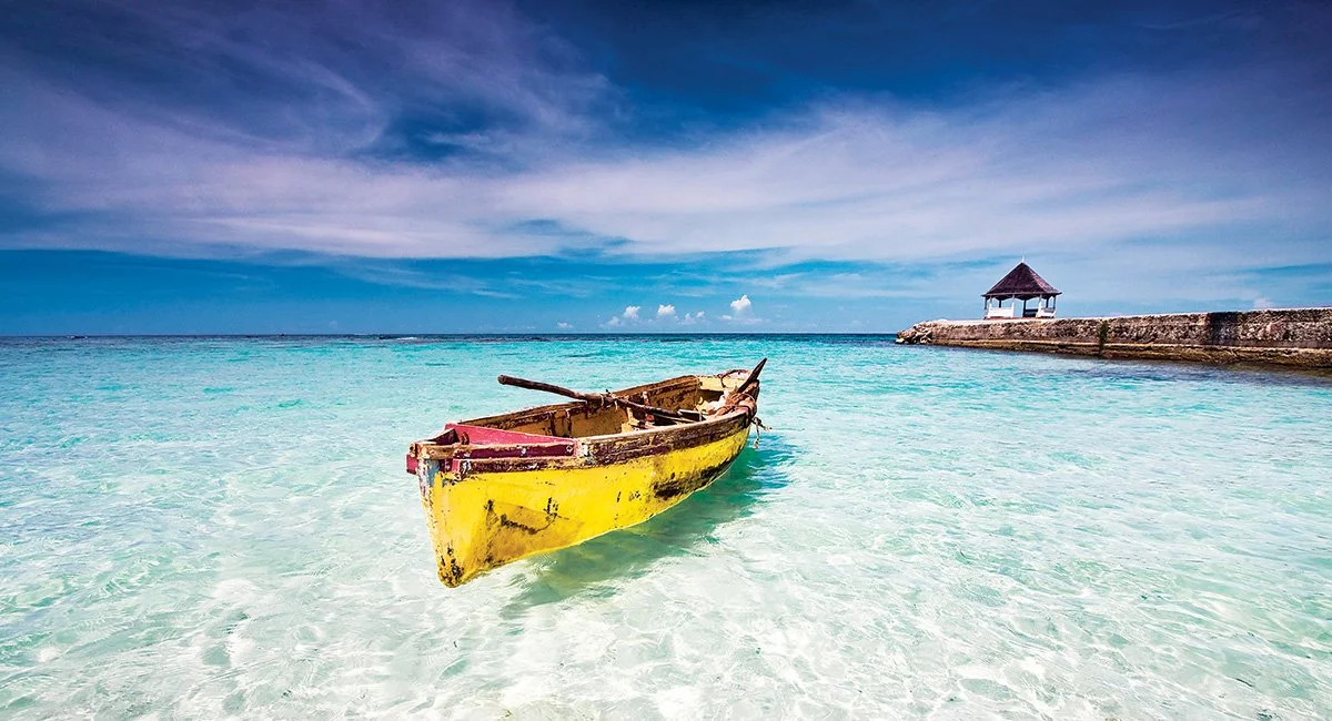 Yellow and red boat floating in clear turquoise water with blue sky and clouds, and a building on the rocky shoreline in the background