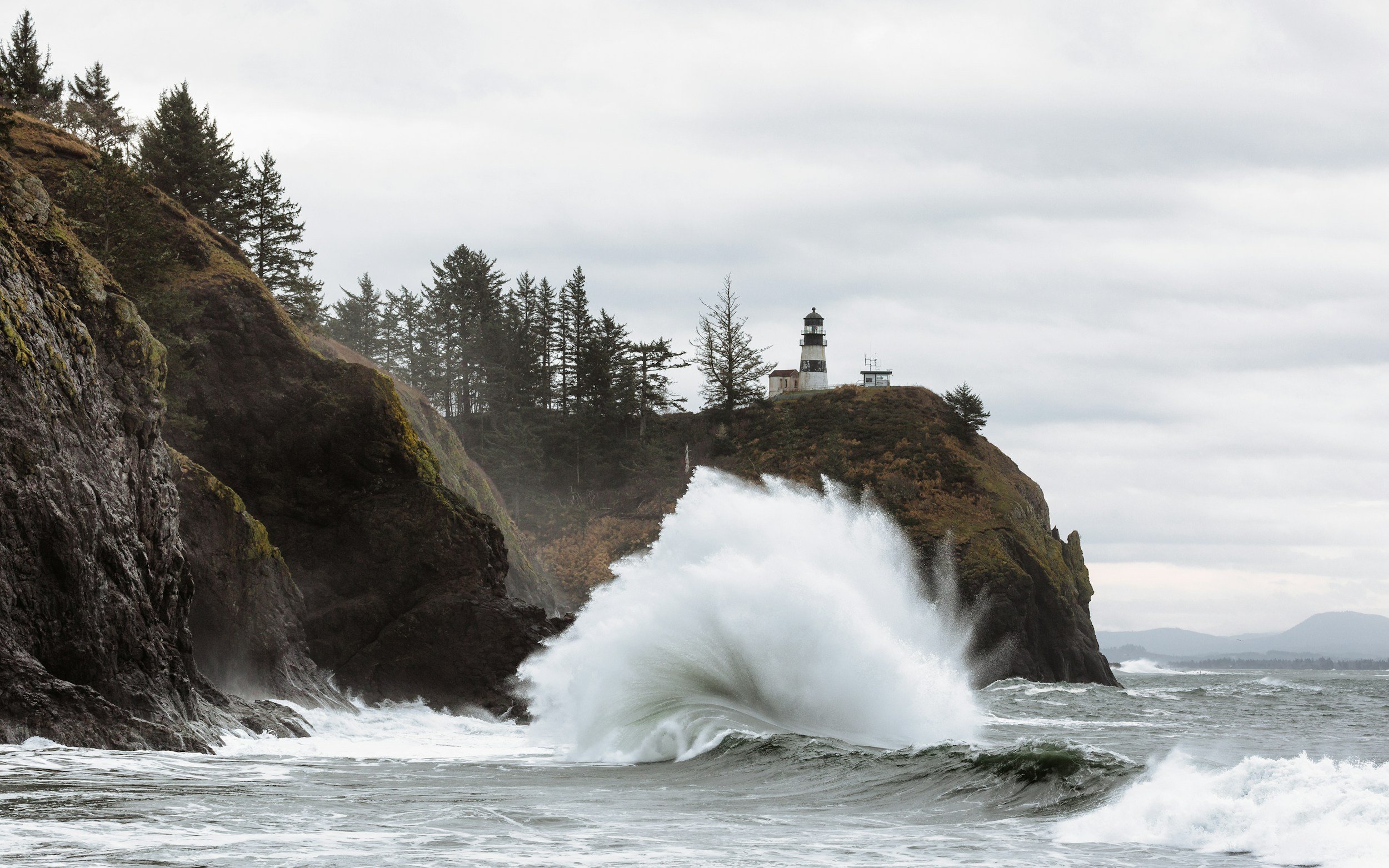 Ocean waves crashing against rocky shoreline with a lighthouse on a hill in the background, cloudy sky.
