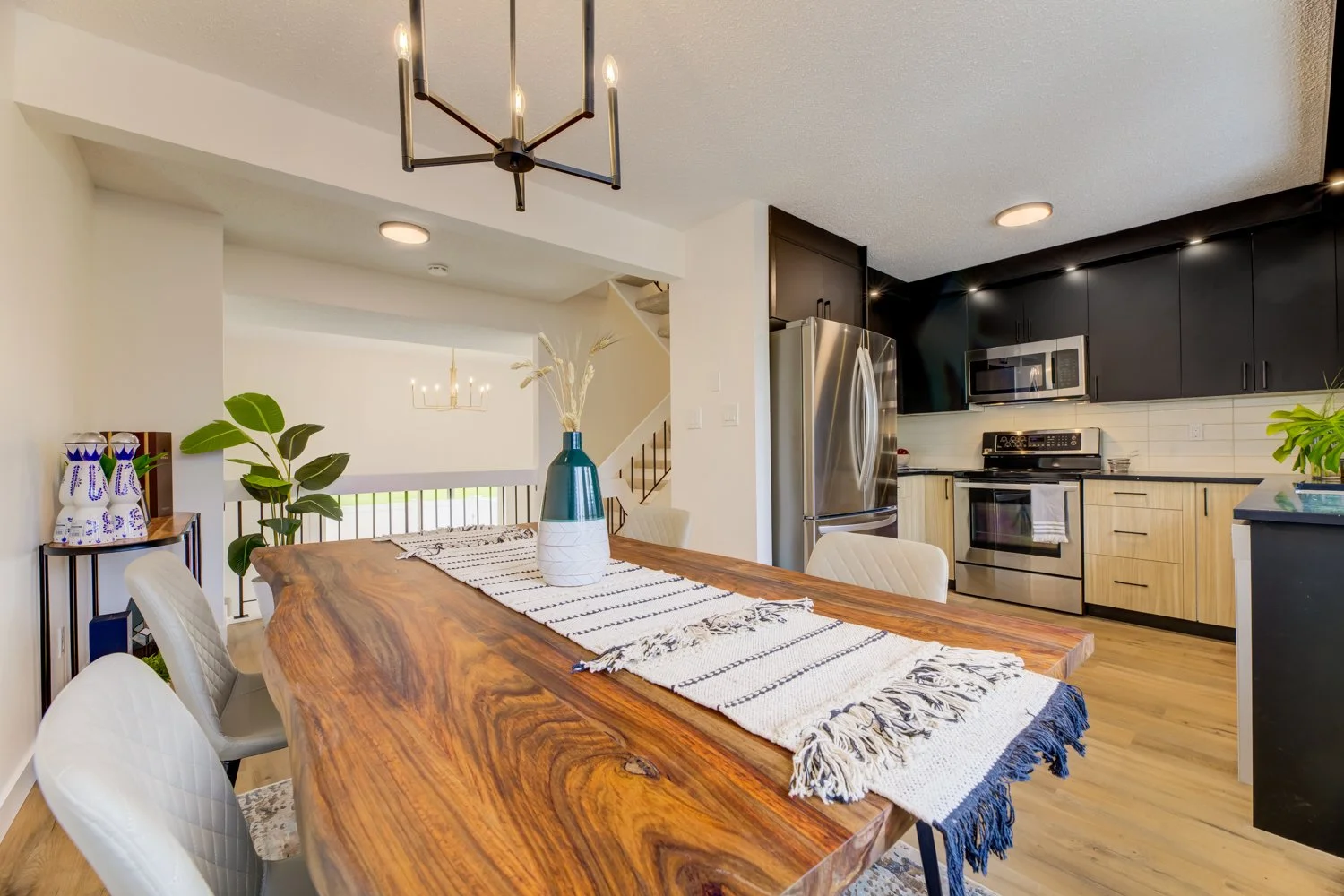 Modern kitchen and dining area with a wooden dining table, white quilted chairs, a decorative vase, black and wooden cabinets, stainless steel appliances, and a staircase in the background.