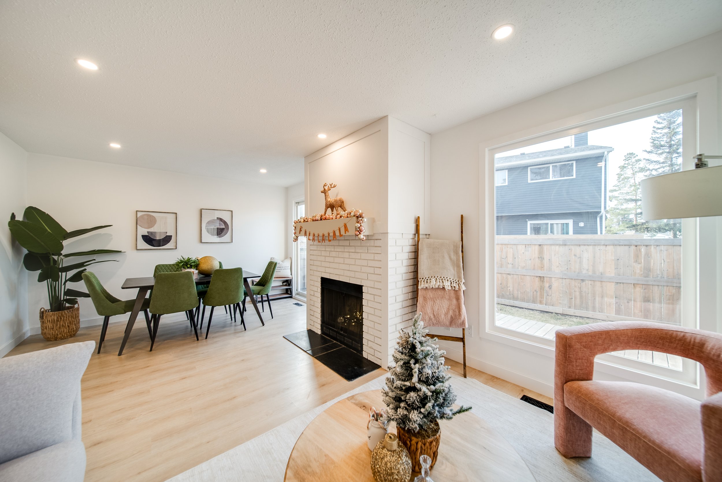 Interior of a living and dining area with a white brick fireplace topped with holiday decorations, a small Christmas tree on a wooden table, and modern furniture including a pink chair and green dining chairs.