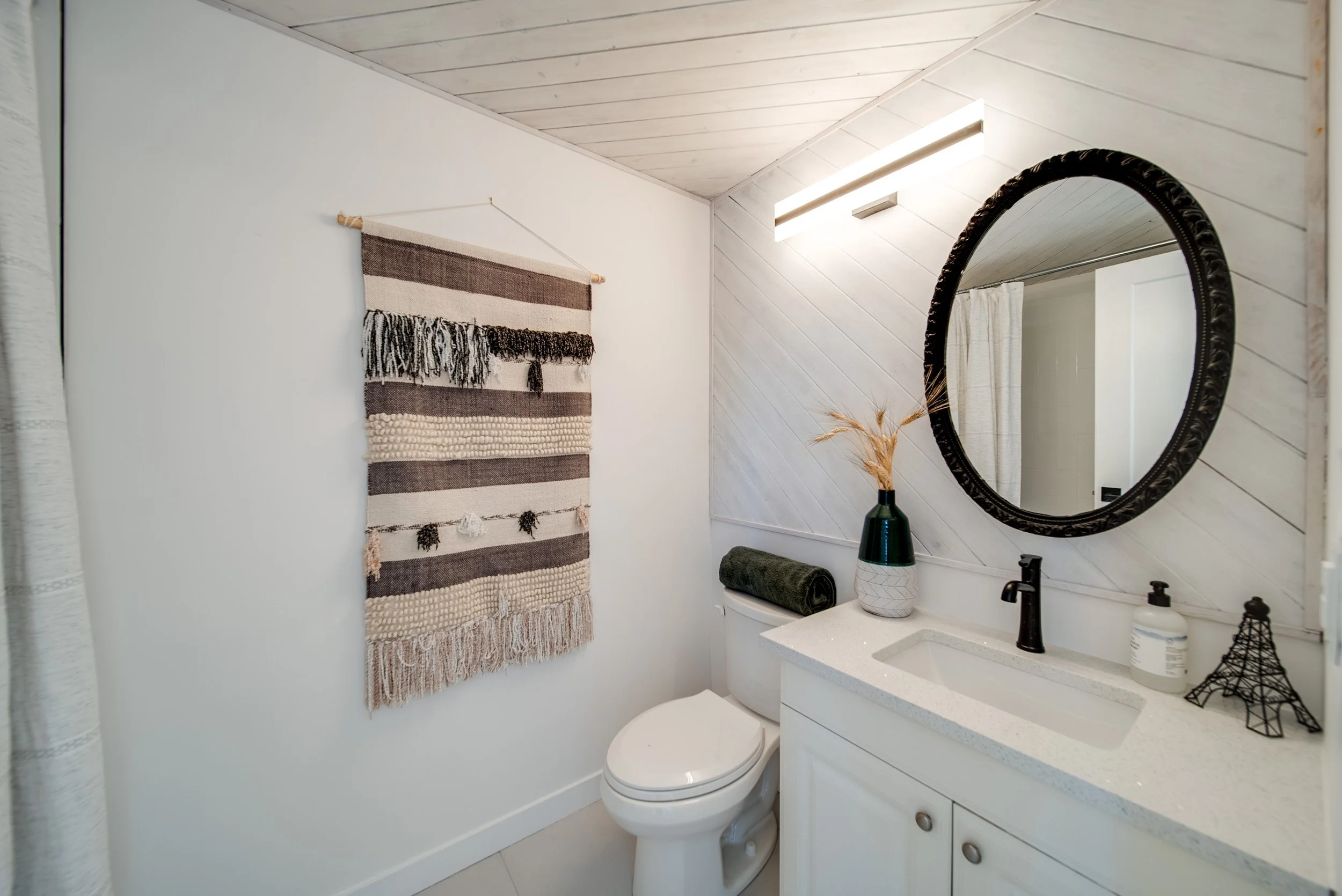 A small bathroom with a white toilet, white vanity with black faucet, a round mirror, and modern decor including a vase with dried grass, a towel, and wall hanging artwork.