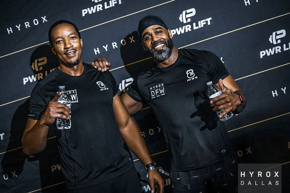 Two men in black athletic shirts holding water bottles, standing in front of a black backdrop with white text and logo that reads 'HYROX DALLAS', 'PWR LIFT', and 'HYROX'. They are smiling, one with hand on the other's shoulder. The man on the right has a beard, headband, and smartwatch.