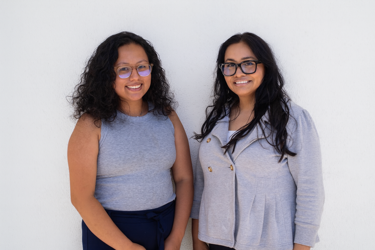 Two women standing side by side smiling in front of a plain white wall.