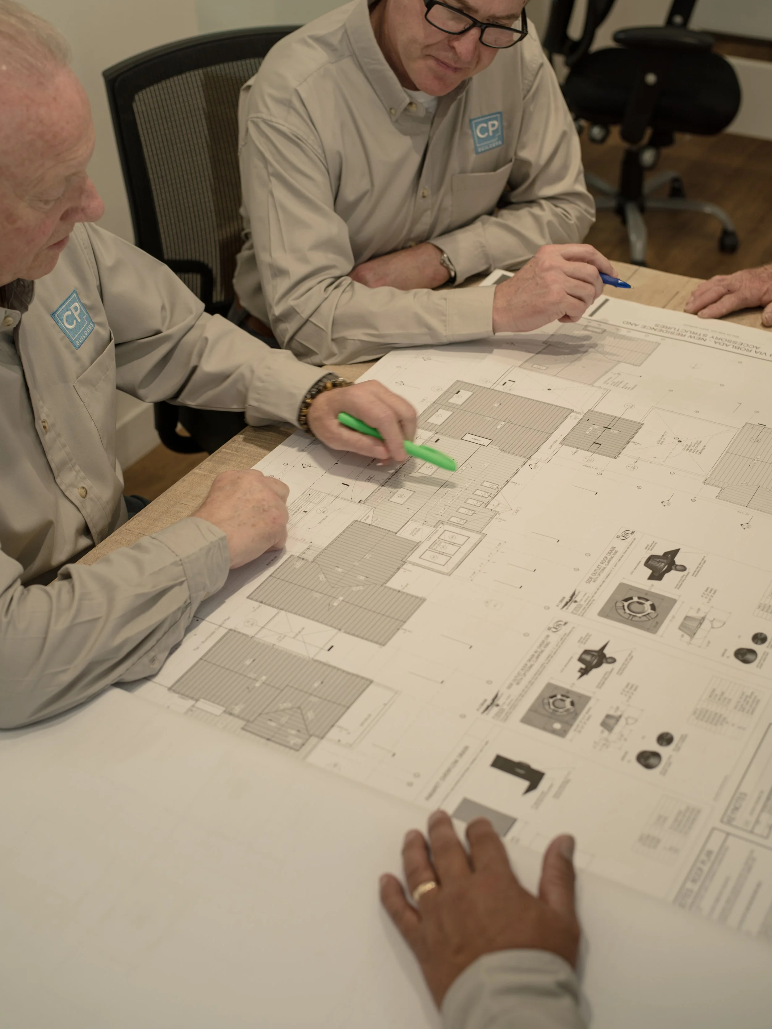 Three men reviewing architectural blueprints at a conference table. Two are holding colored markers, discussing the plans.
