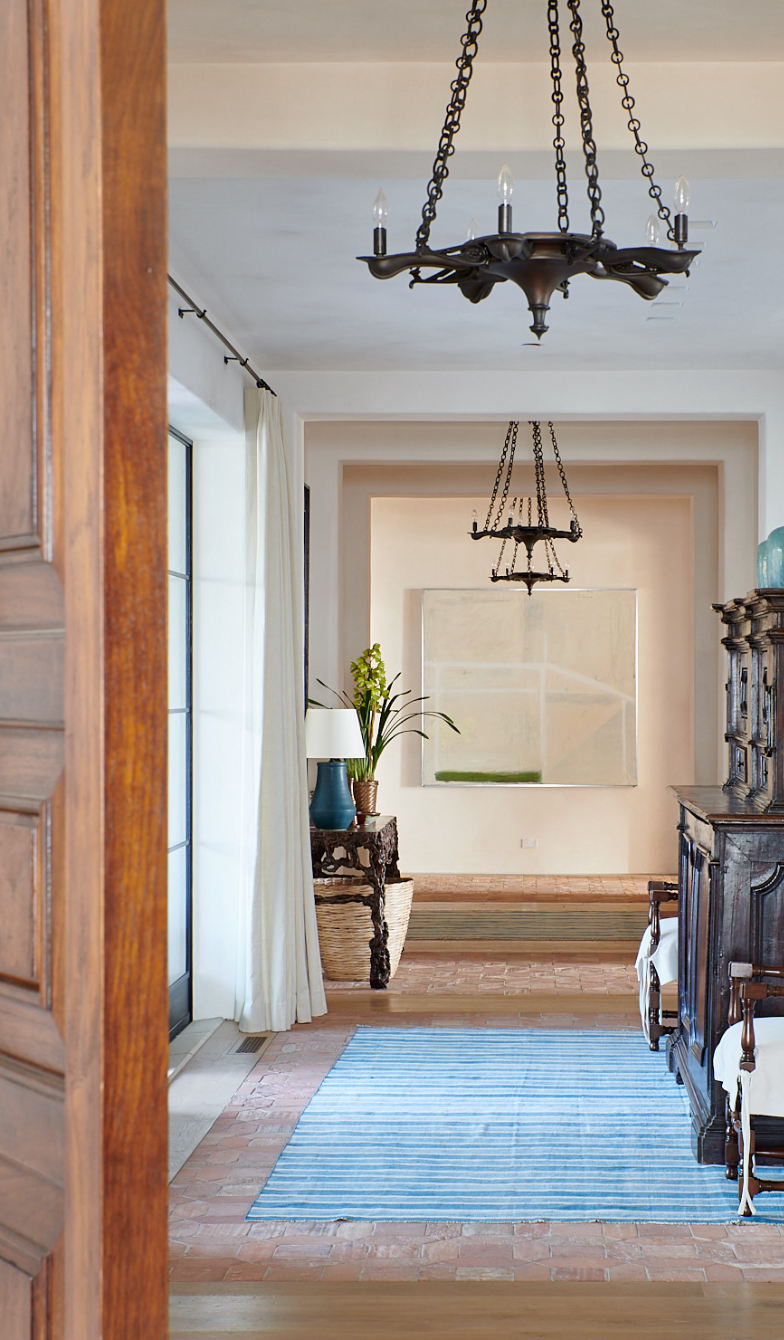 Interior view of a living space with a brick floor, wooden furniture, a blue and white rug, a hanging black chandelier, a window with white curtains, and decorative items.