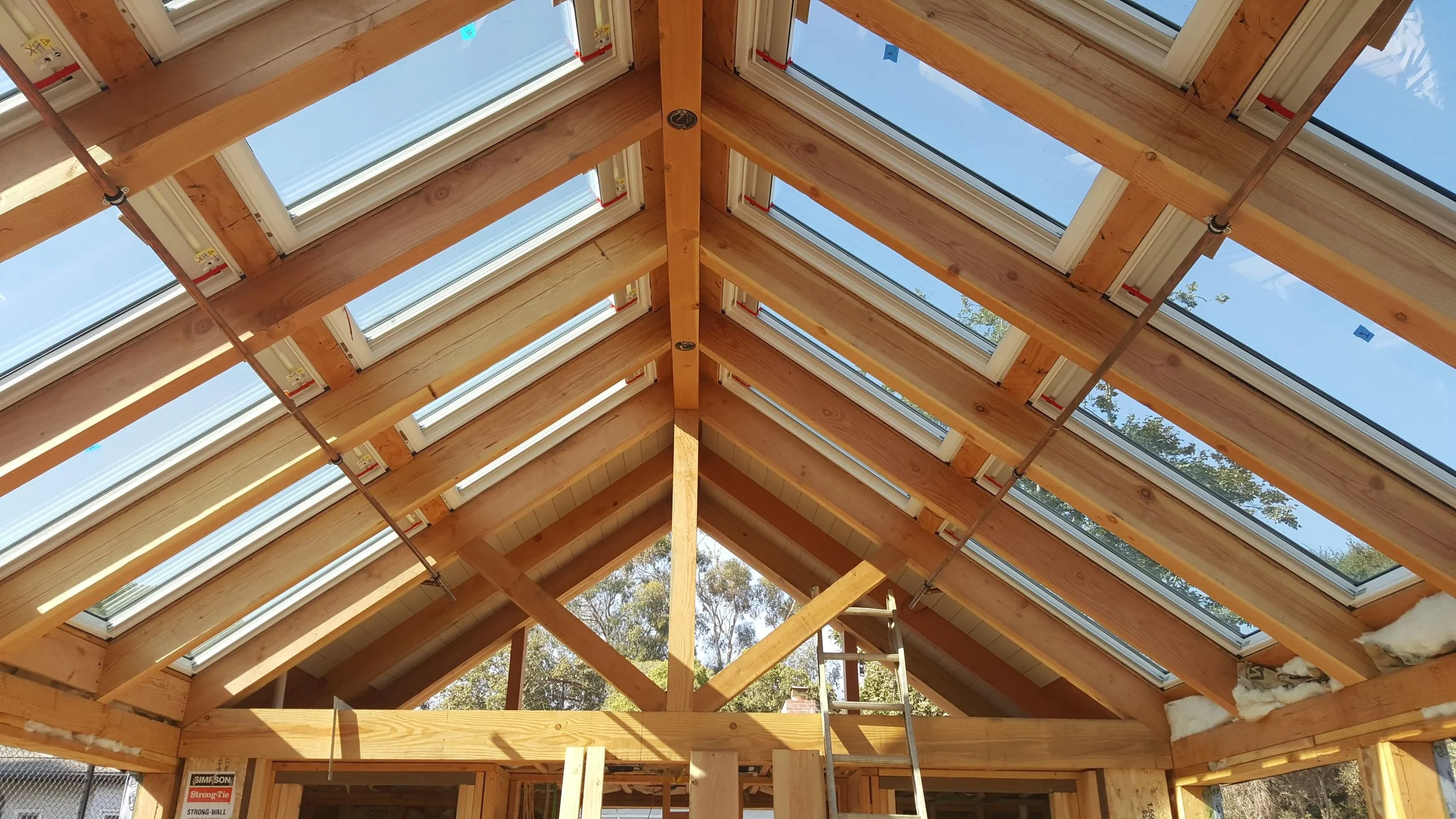 Inside view of a building under construction showing wooden roof framing with multiple skylights and a ladder.