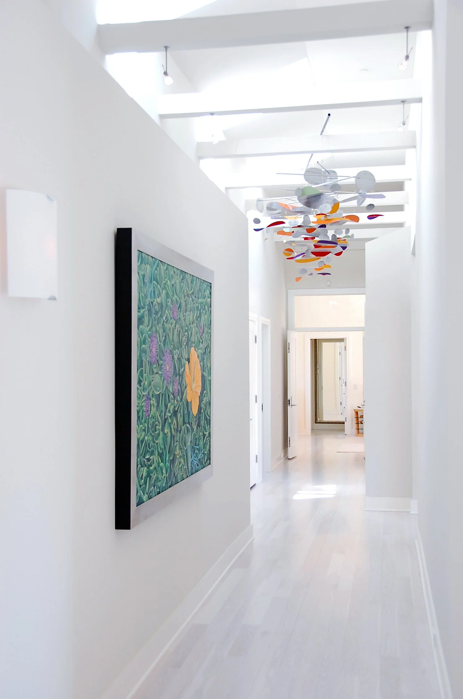 A bright, minimalist hallway with white walls, a colorful abstract hanging sculpture on the ceiling, and a framed painting of green foliage and flowers on the wall.