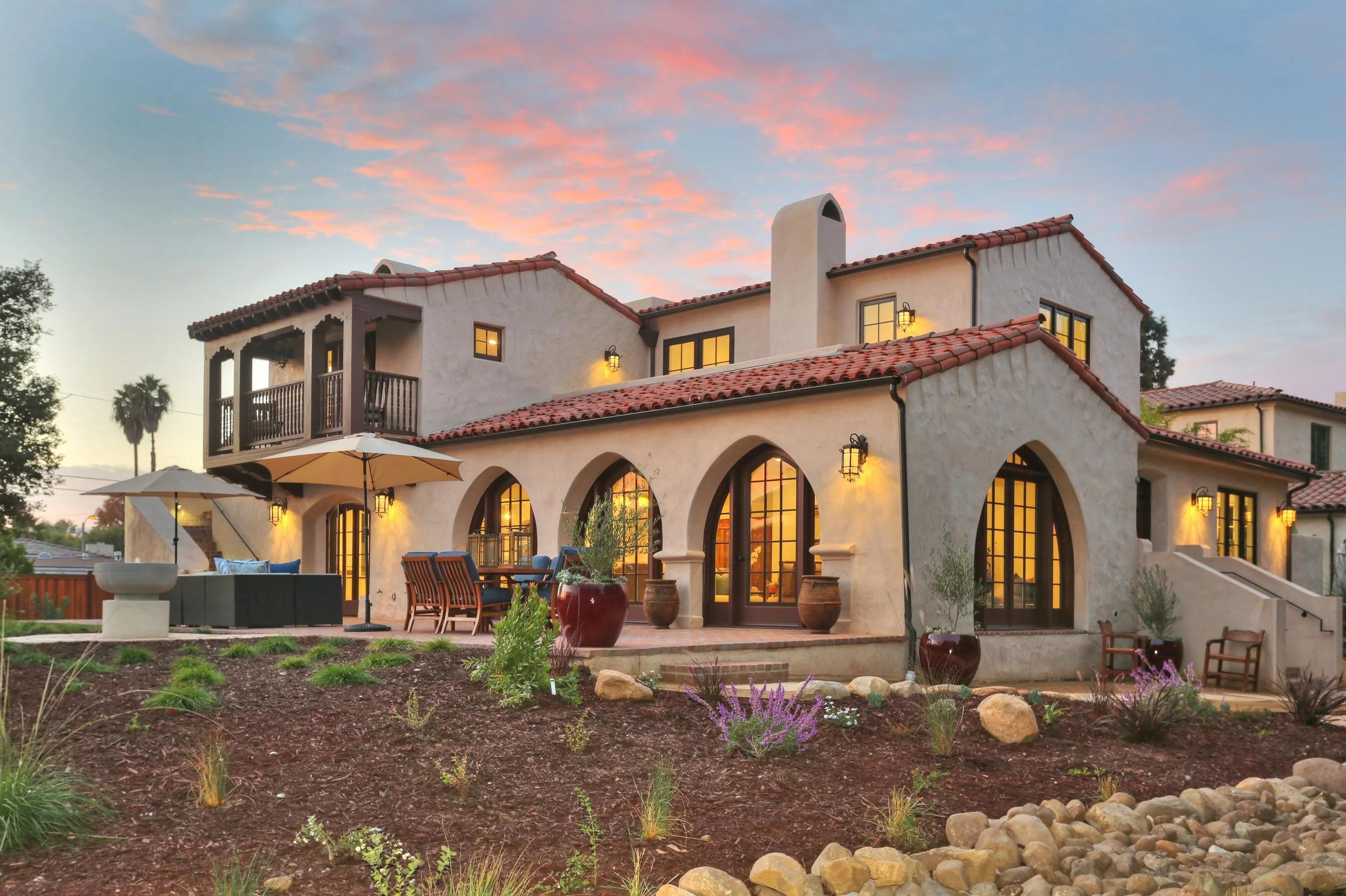 A two-story Mediterranean-style house with a stucco exterior, red tile roof, and arched windows and doors. The house features a balcony, outdoor seating area with umbrellas, potted plants, landscaping with rocks, and is illuminated by outdoor lights at sunset.