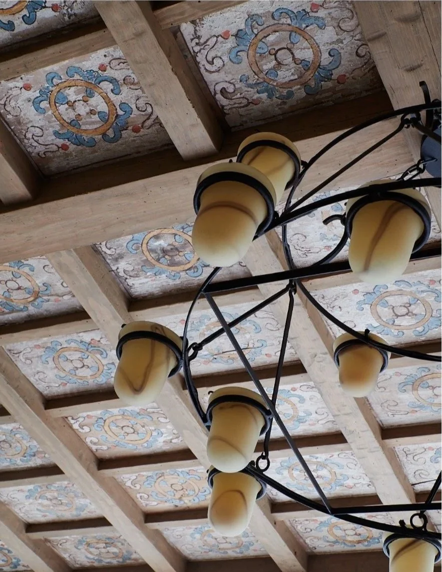 Chandelier with multiple yellowish glass shades hanging from a decorative wooden ceiling with painted patterns.