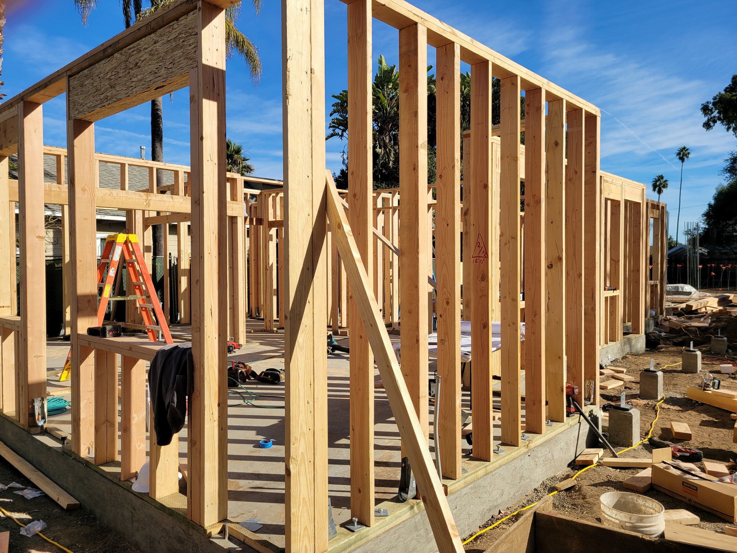 Wooden framing construction of a building with a blue sky, a few palm trees, and construction tools and materials nearby.