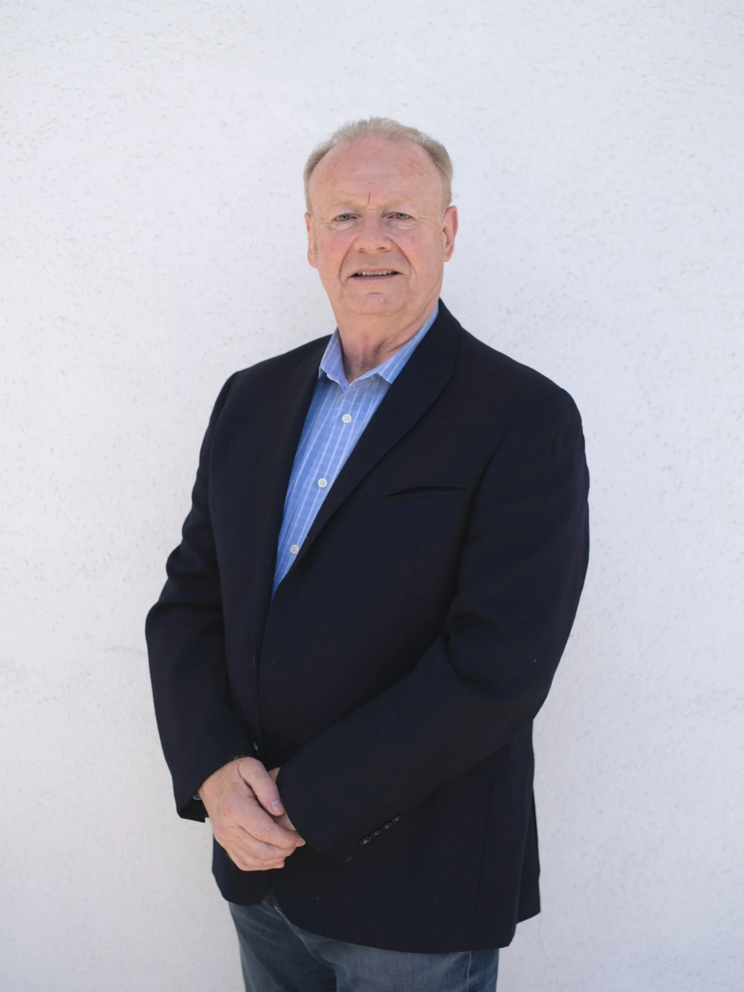 Gavin, the contractor and co-founder, wearing a black blazer and a blue checkered shirt standing against a white textured wall.