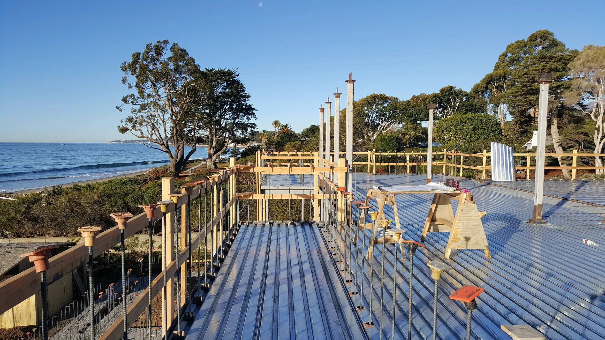 Construction site on the beach with wooden framing and metal roofing, overlooking the ocean with trees in the background.