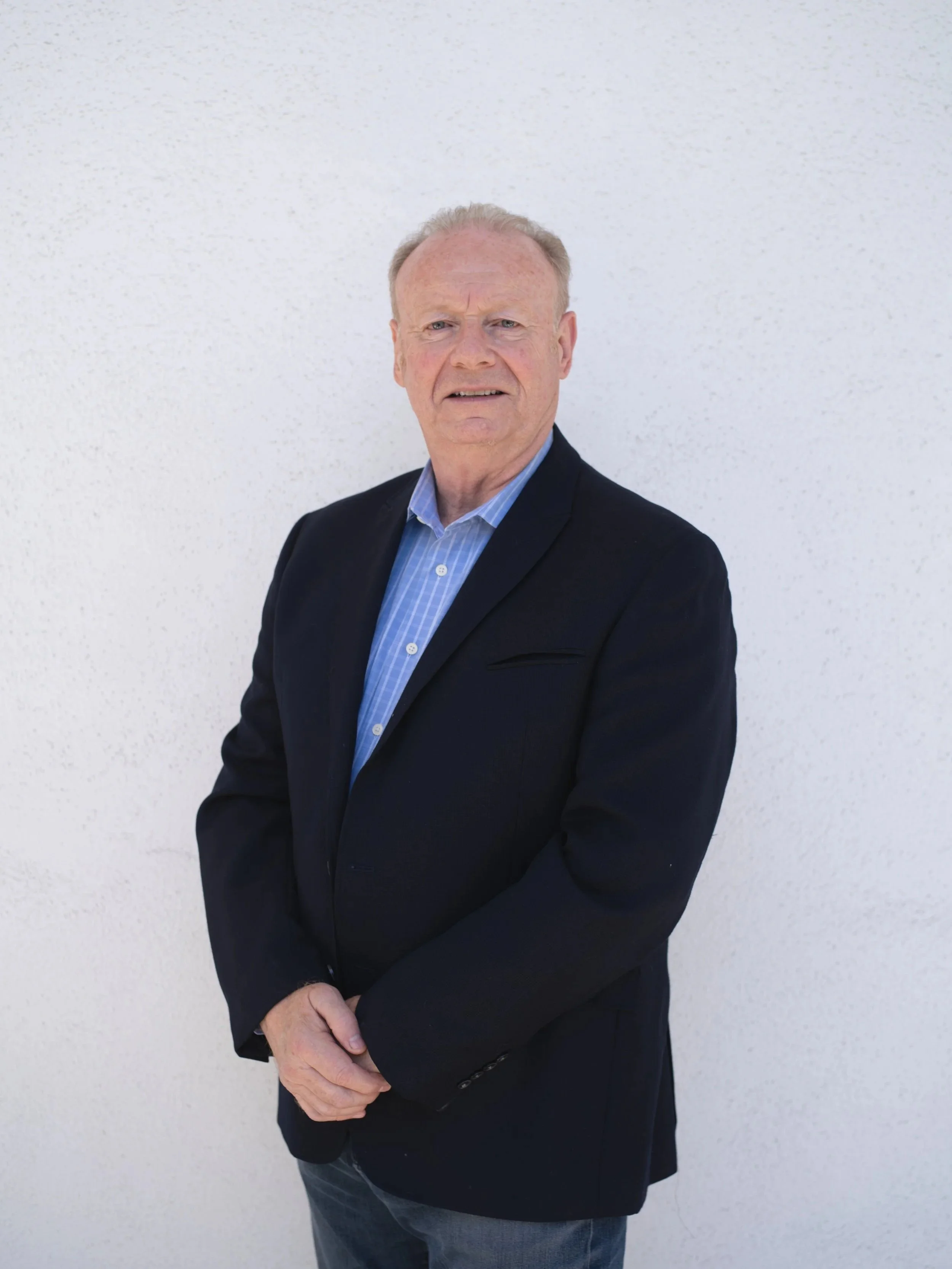 Gavin, the contractor and co-founder, wearing a black blazer and a blue checkered shirt standing against a white textured wall.
