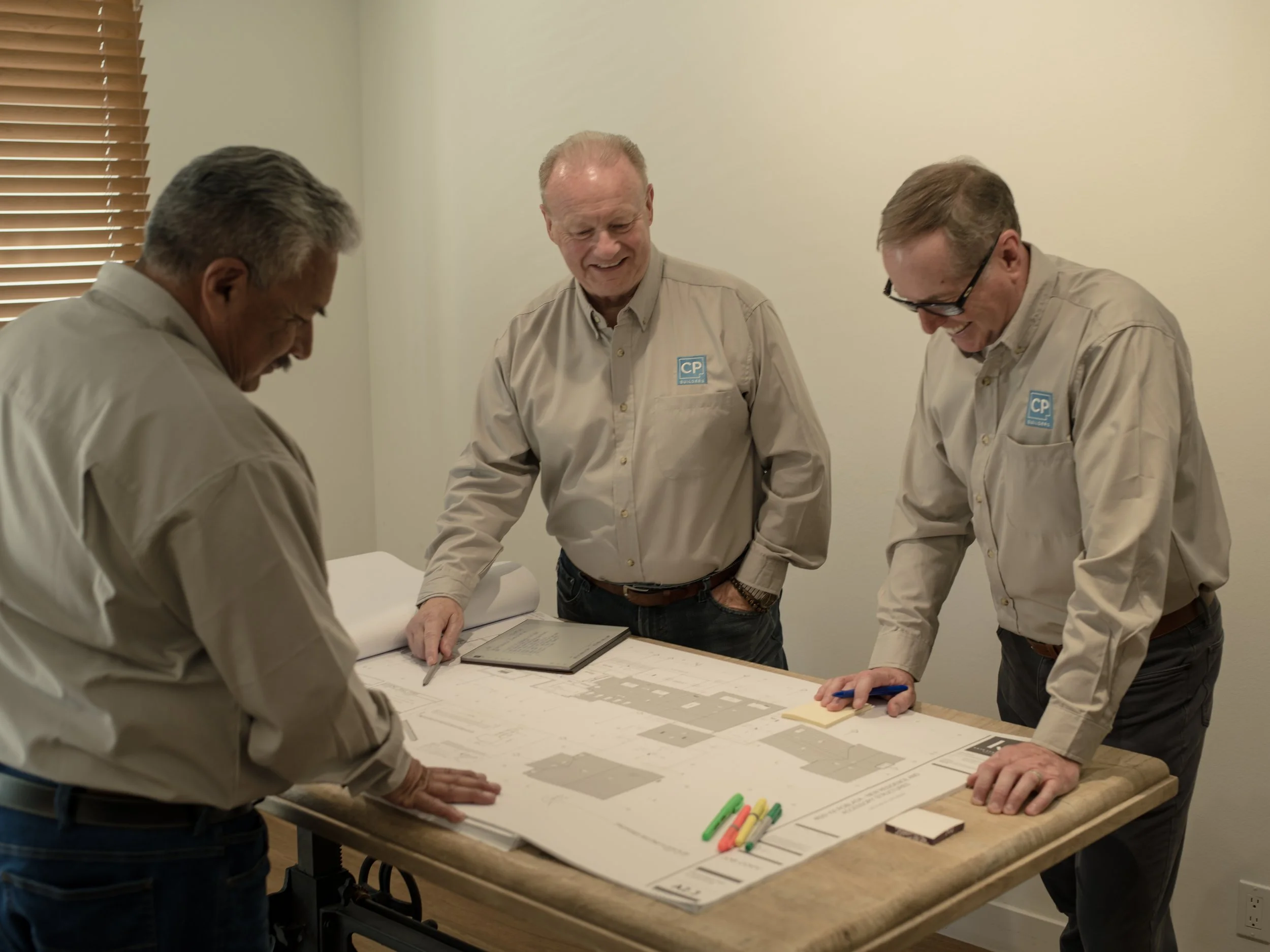 Three men in beige shirts gathered around a table examining a large blueprint or architectural plan, smiling and engaged in discussion.