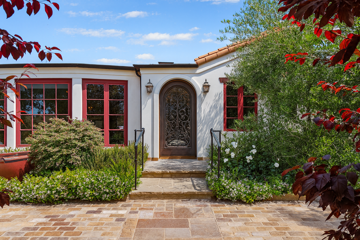 Front entrance of a house with a decorative black metal door, surrounded by greenery and white flowers. The house has white walls with red window frames, and there are two lantern-style lights on either side of the door. A brick pathway leads up to the step in front of the door.