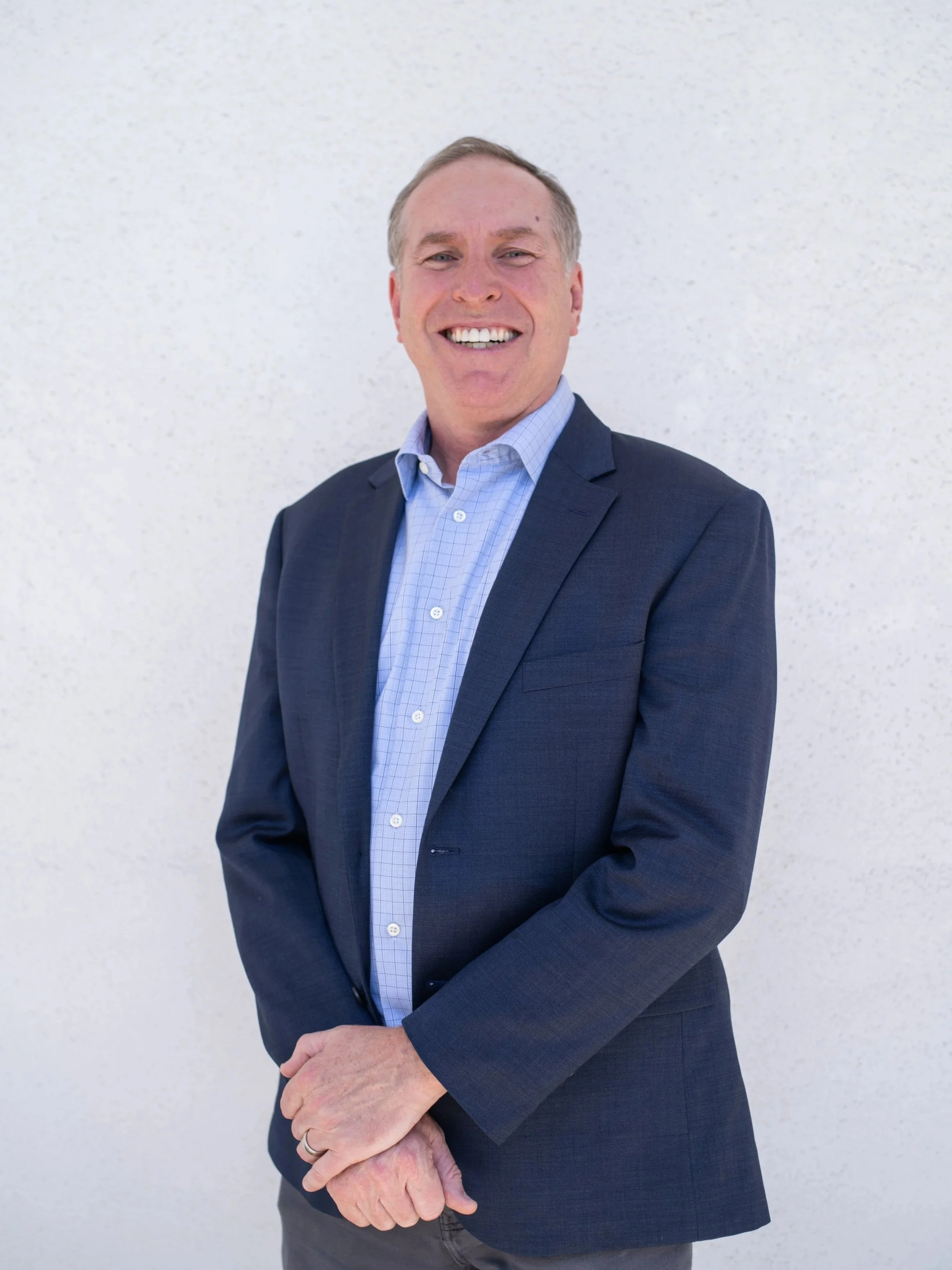 Michael, a smiling middle-aged man wearing a dark blue suit jacket and a light blue checkered dress shirt, standing in front of a plain white wall.